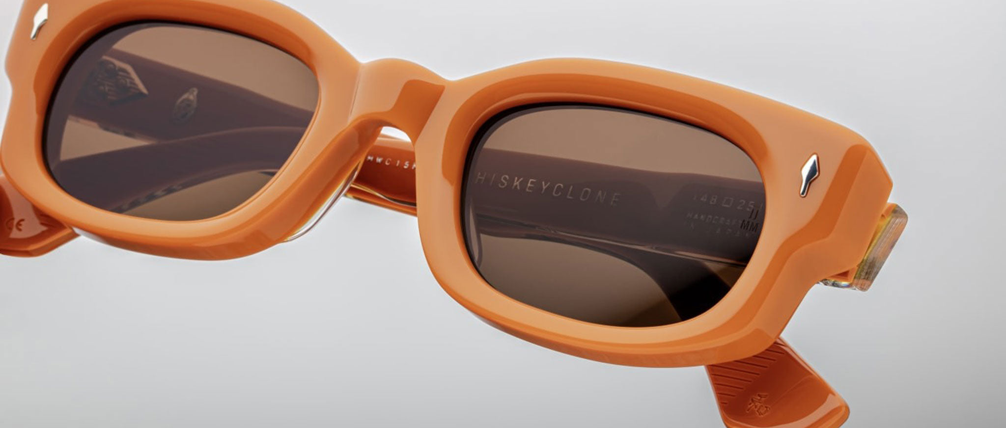 Close-up of Jacques Marie Mage Whiskeyclone sunglasses in orange with brown lenses, highlighting thick frames, hinge detail, and WHISKEYCLONE engraving on the inner arm. Set against a neutral gray background.