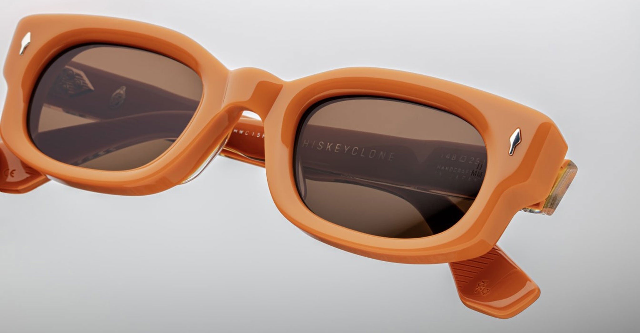 Close-up of Jacques Marie Mage Whiskeyclone sunglasses in orange with brown lenses, highlighting thick frames, hinge detail, and WHISKEYCLONE engraving on the inner arm. Set against a neutral gray background.