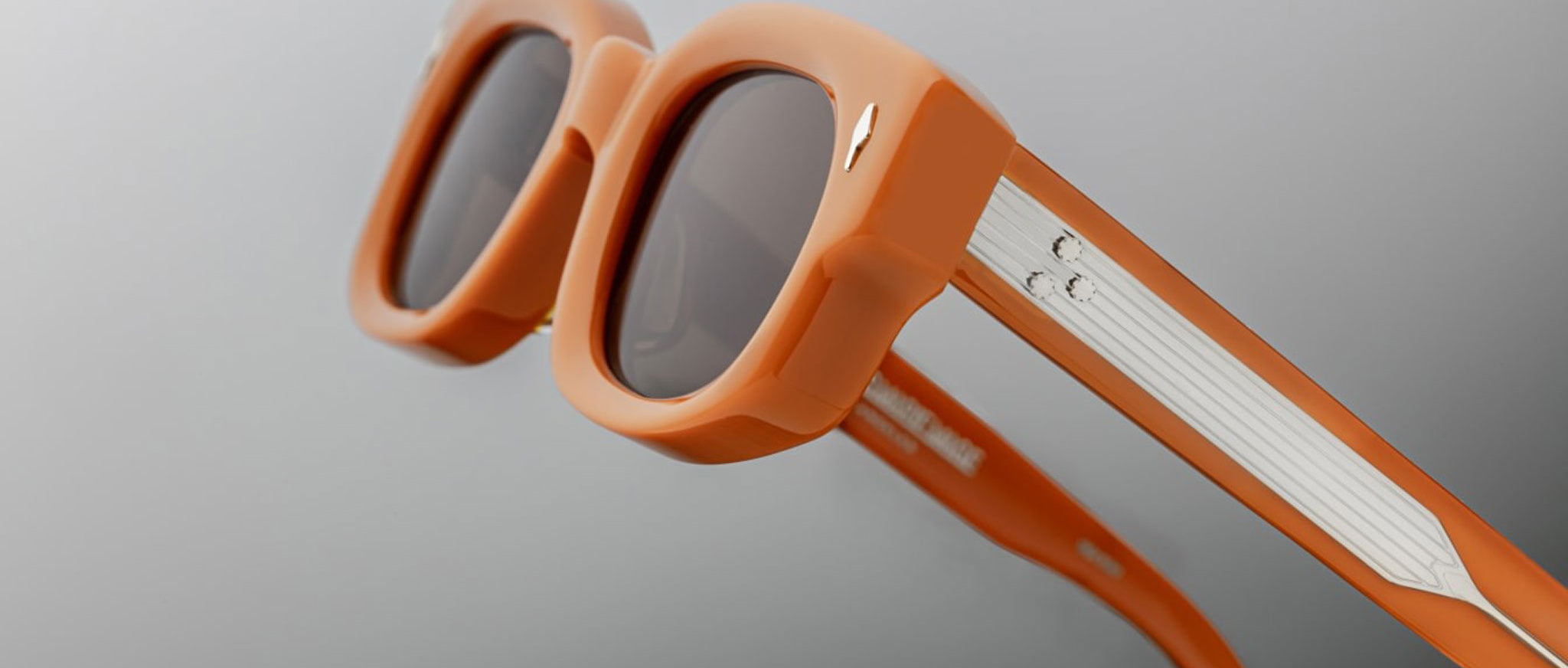Close-up view of Jacques Marie Mage Whiskeyclone orange rectangular sunglasses with dark lenses and metallic accents on the arms, set against a plain light gray background.