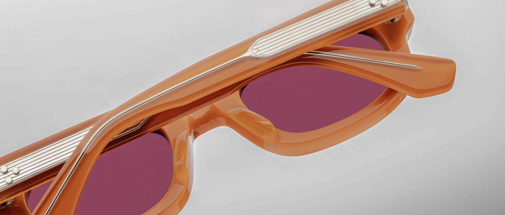 Close-up of Jacques Marie Mage Whiskeyclone sunglasses with orange rectangular frames and red-tinted lenses, showing top and side details against a light background.