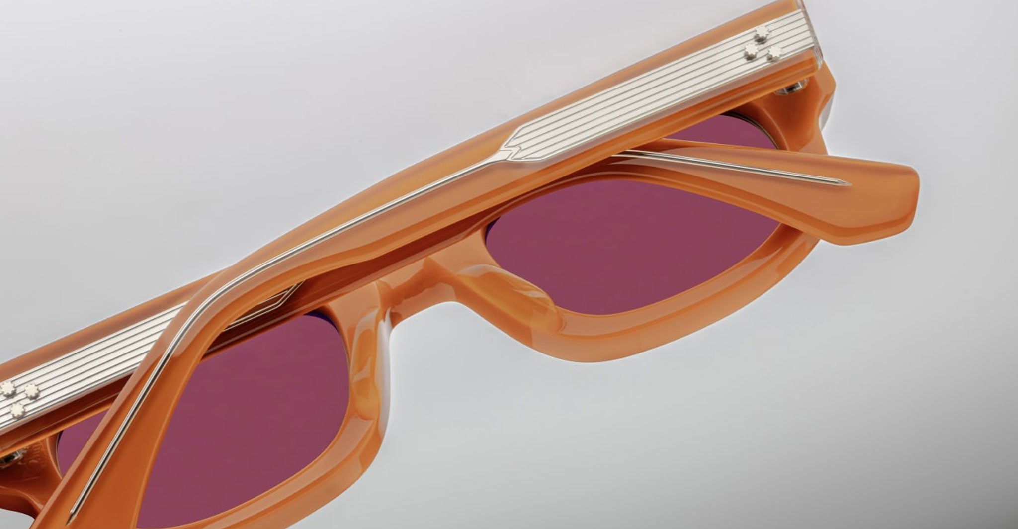 Close-up of Jacques Marie Mage Whiskeyclone sunglasses with orange rectangular frames and red-tinted lenses, showing top and side details against a light background.