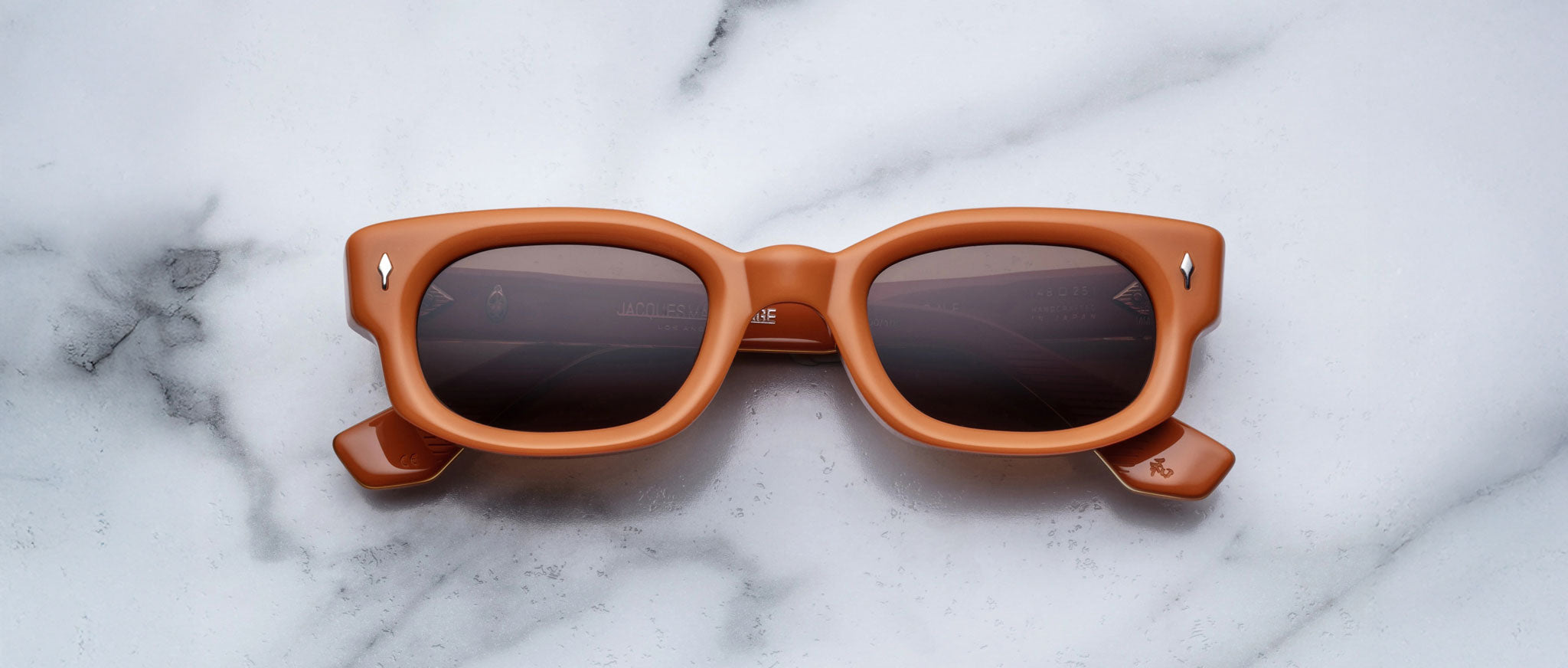 The Jacques Marie Mage Whiskeyclone sunglasses feature square-shaped, thick light brown frames with dark lenses, displayed on a white marble surface with gray veining.