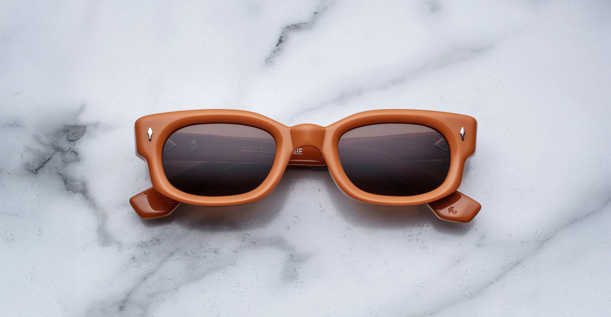 The Jacques Marie Mage Whiskeyclone sunglasses feature square-shaped, thick light brown frames with dark lenses, displayed on a white marble surface with gray veining.