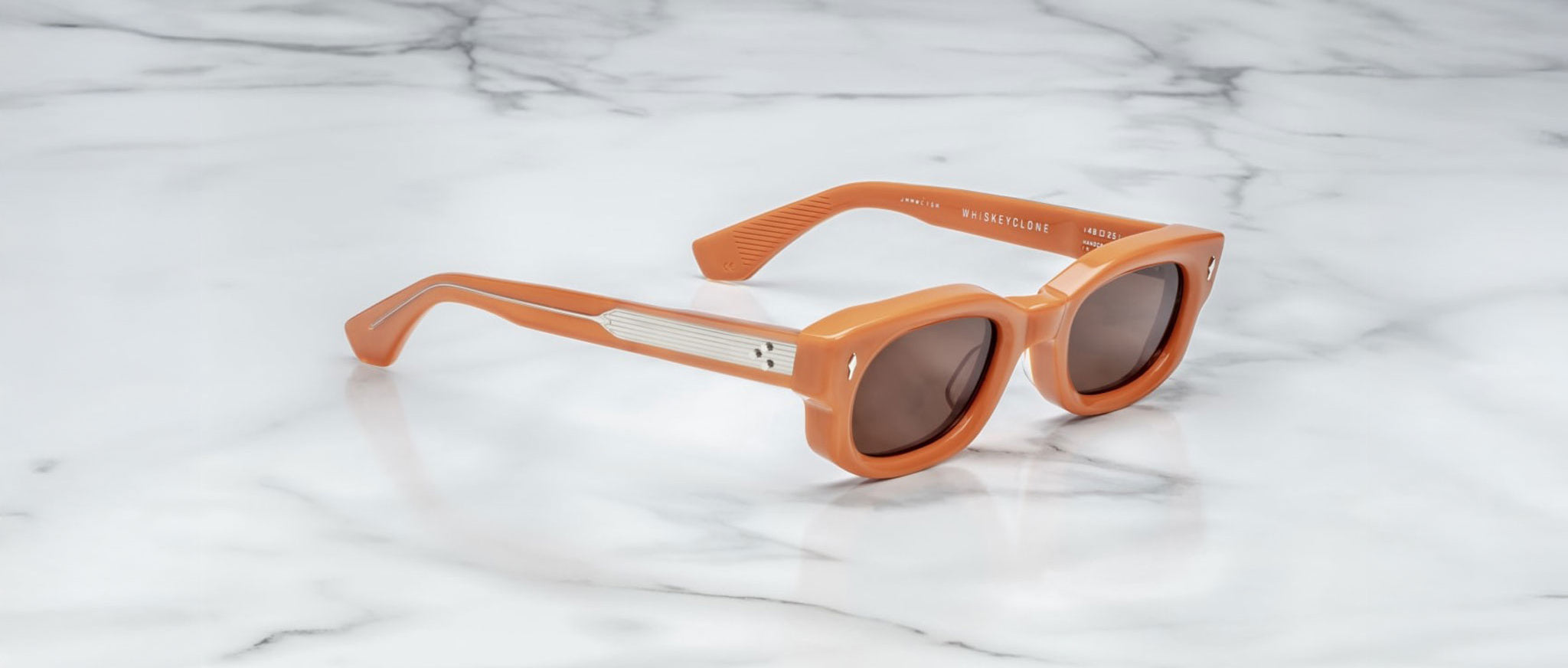 A pair of Jacques Marie Mage Whiskeyclone orange rectangular sunglasses with dark lenses and silver arm detailing, placed on a white marble surface.
