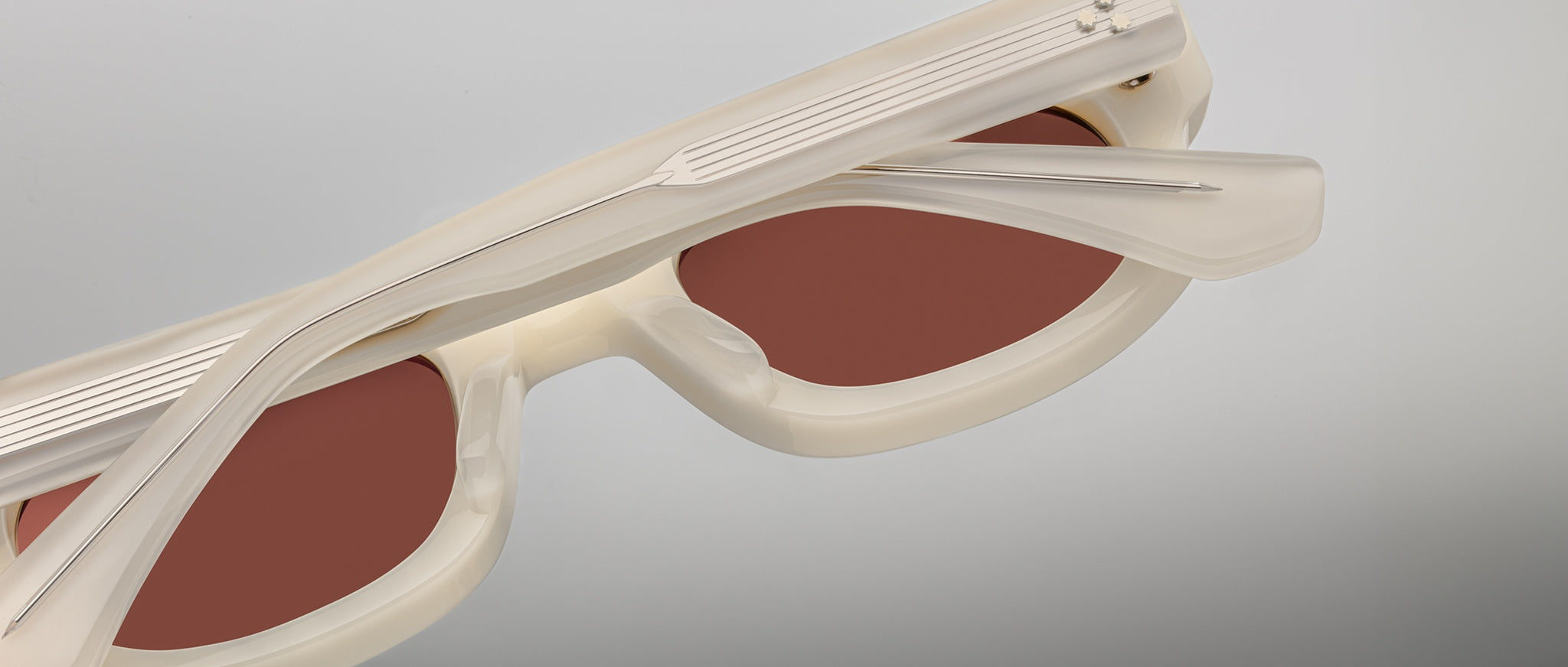 A close-up of Jacques Marie Mage Whiskeyclone sunglasses, featuring a cream-colored translucent frame, brown lenses, metallic hinges, and a minimalist modern design, shown folded against a plain background.