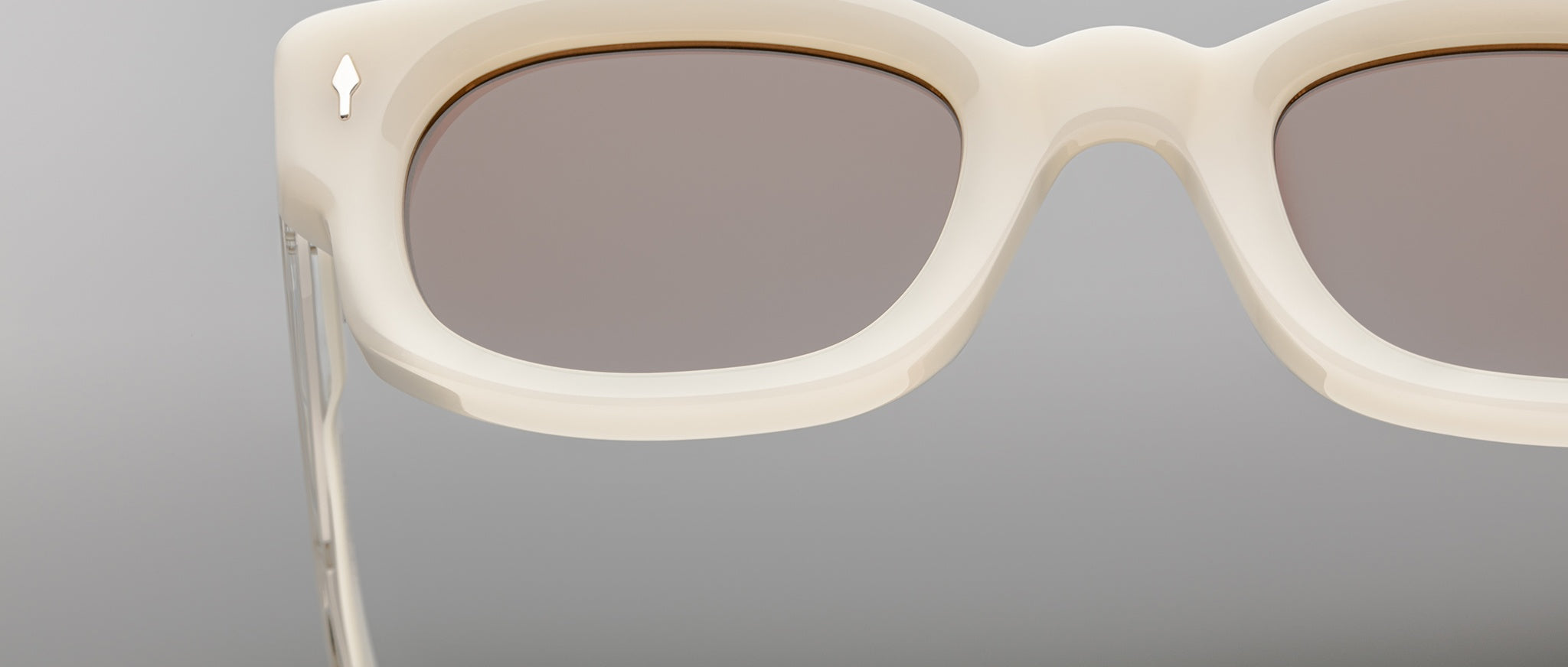 Close-up view of Jacques Marie Mage Whiskeyclone sunglasses featuring thick off-white rims, light brown lenses, and a metallic arrow detail on the temple, set against a neutral gray background.