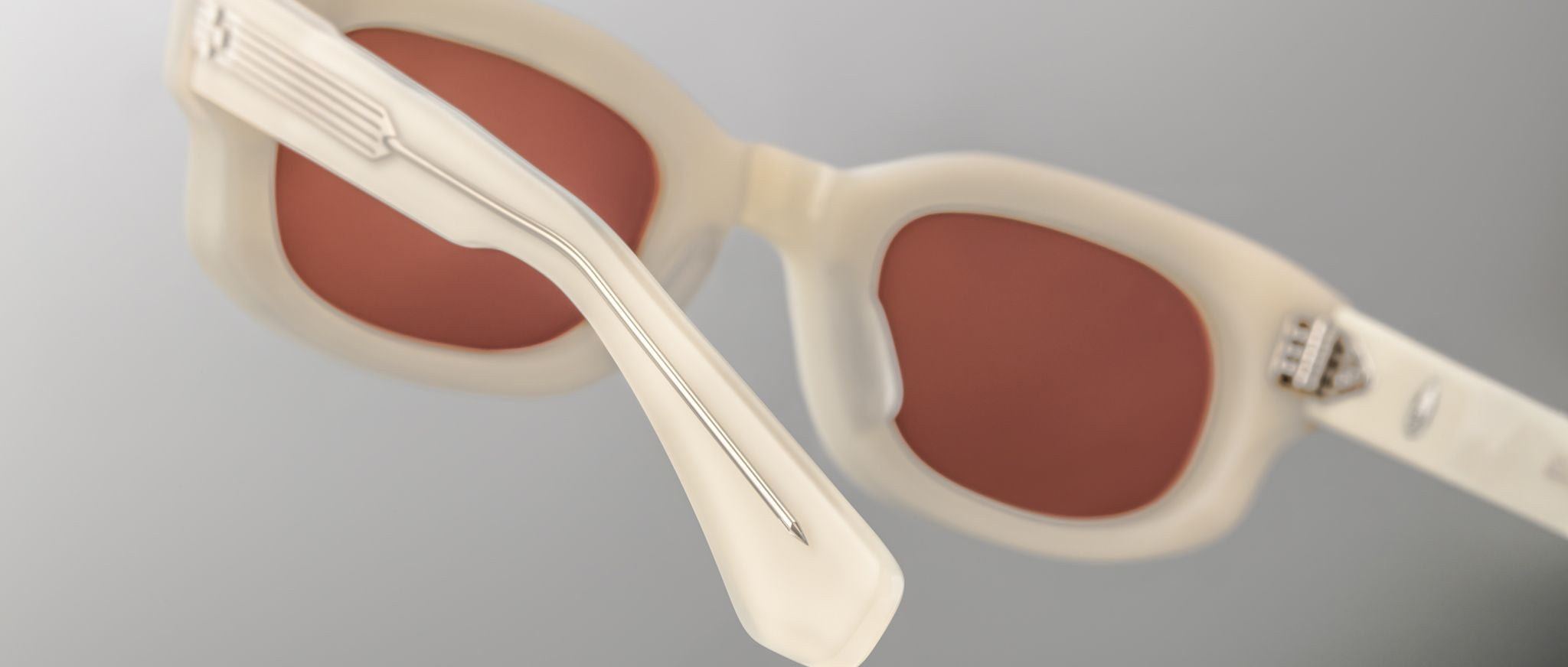 Close-up of Jacques Marie Mage Whiskeyclone sunglasses featuring thick, off-white frames and reddish-brown lenses, set against a gray gradient background. The image emphasizes the detailed temples and lens craftsmanship.