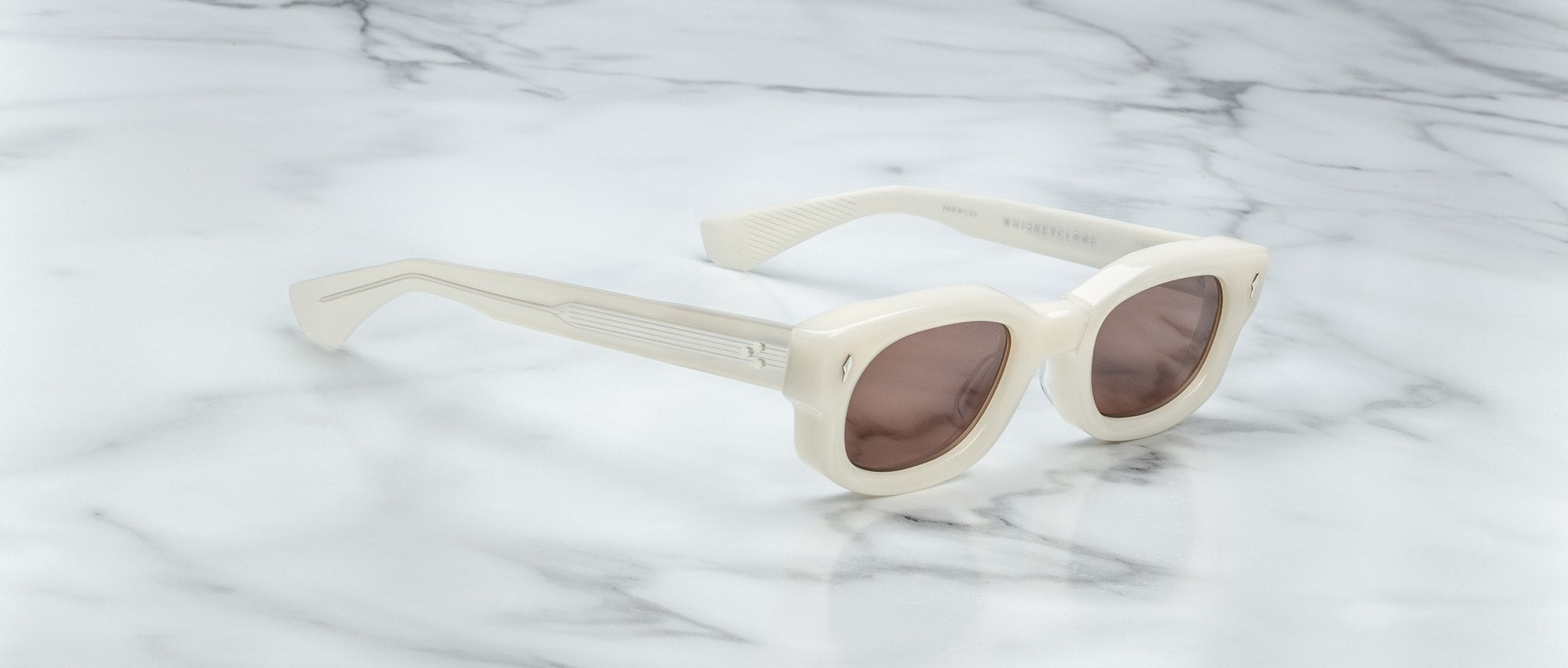 Jacques Marie Mage Whiskeyclone sunglasses with cream-colored, round tinted lenses and thick frames rest on a white marble surface.