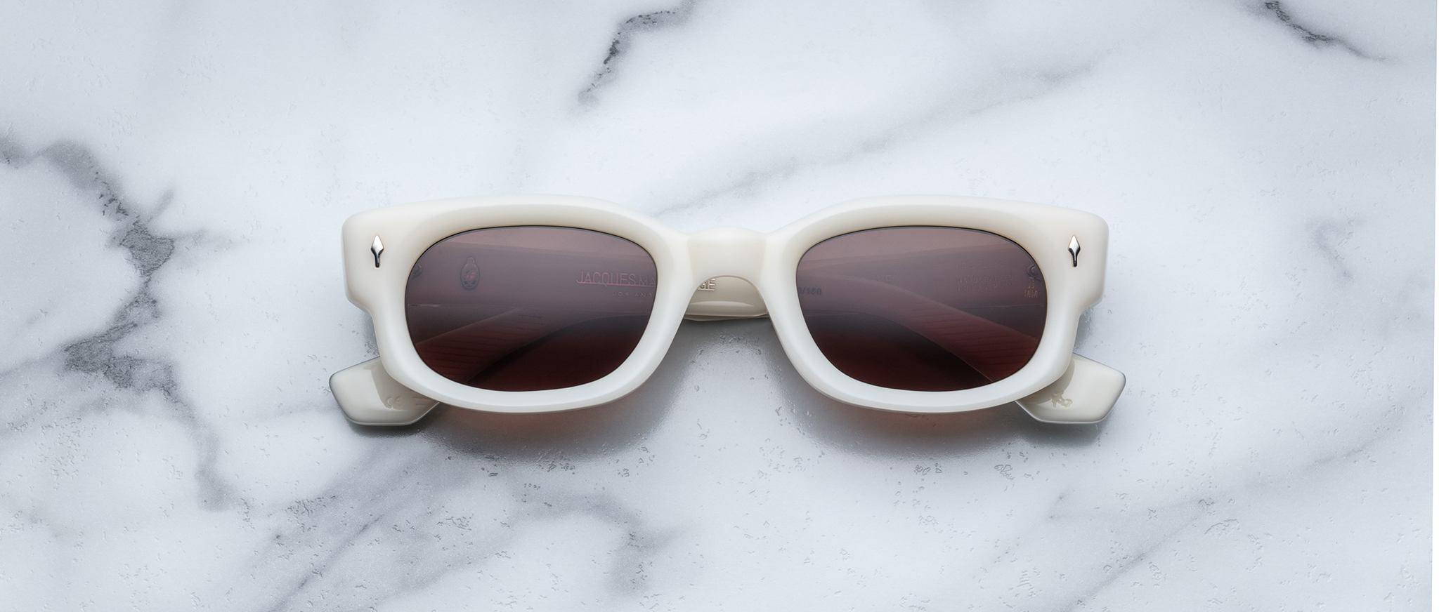 Jacques Marie Mage Whiskeyclone sunglasses with white frames and dark lenses are showcased on a white marble surface with grey veining.