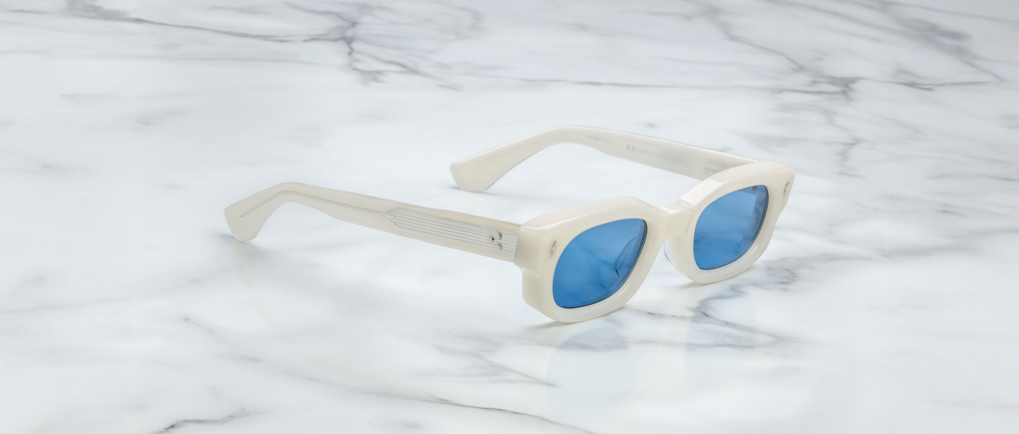 The Jacques Marie Mage Whiskeyclone sunglasses, featuring white frames and blue-tinted lenses, rest on a white marble surface with gray veins.