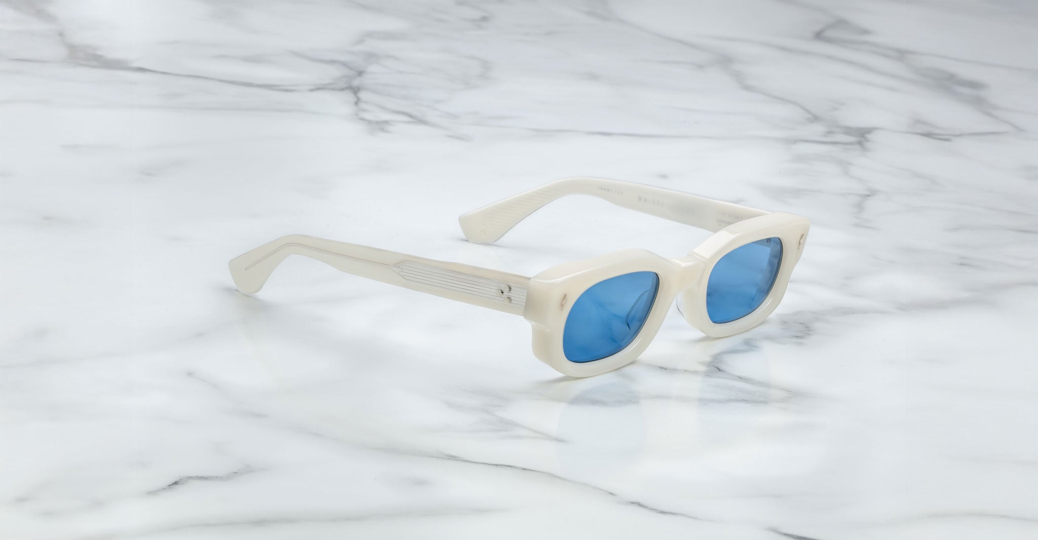 The Jacques Marie Mage Whiskeyclone sunglasses, featuring white frames and blue-tinted lenses, rest on a white marble surface with gray veins.