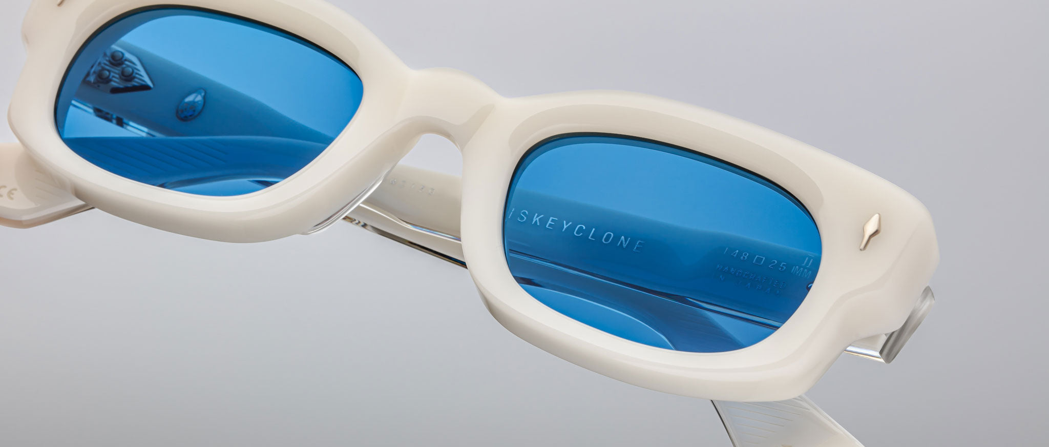 Close-up of white-framed Jacques Marie Mage Whiskeyclone sunglasses with bright blue tinted lenses, reflecting light and featuring small metallic details. The brand name is visible on the inside of the left lens.