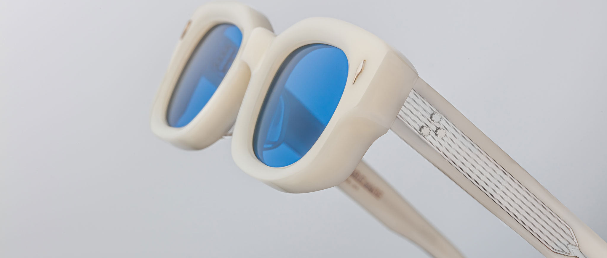 Close-up of Jacques Marie Mage Whiskeyclone sunglasses with thick cream frames, blue-tinted lenses, silver striped arms, and a small decorative detail, set against a light gray background.