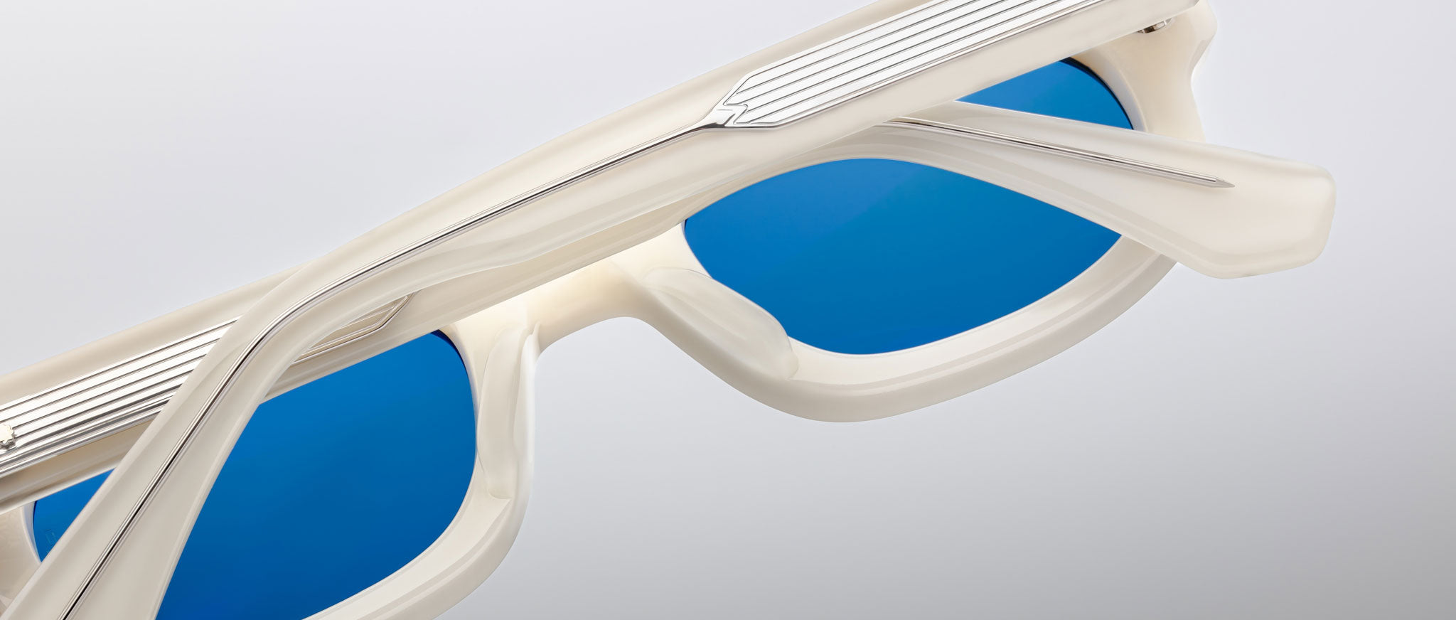 Close-up of Jacques Marie Mage Whiskeyclone sunglasses with white frames and blue lenses, viewed at an angle from above. Partially folded arms display metal accents against a plain light gray background.