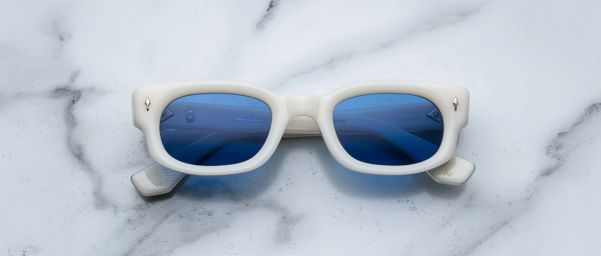 Jacques Marie Mage Whiskeyclone sunglasses, featuring a rectangular white frame and blue lenses, rest facing upward on a white marble surface, with the lenses reflecting hints of the surrounding environment.