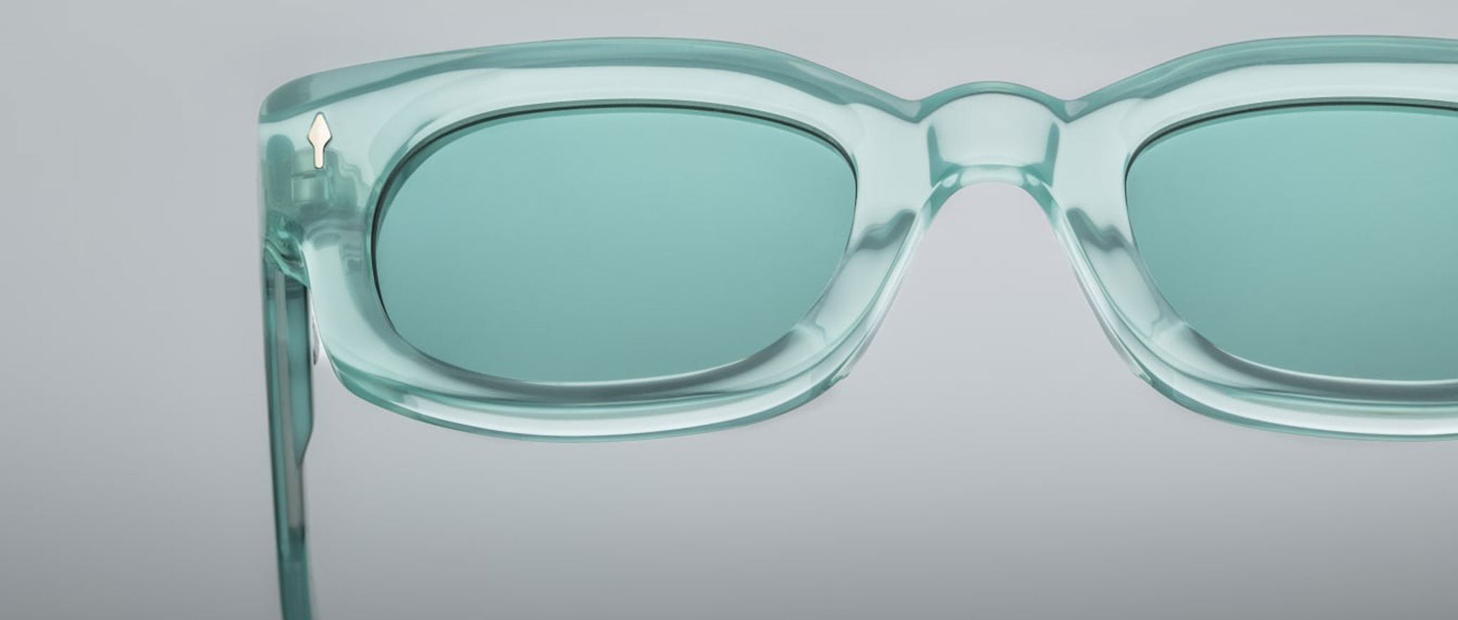 Close-up of Jacques Marie Mage Whiskeyclone sunglasses in translucent teal, featuring rectangular frames and matching tinted lenses. Set on a plain light gray background with only the top half and right side of the frame visible.