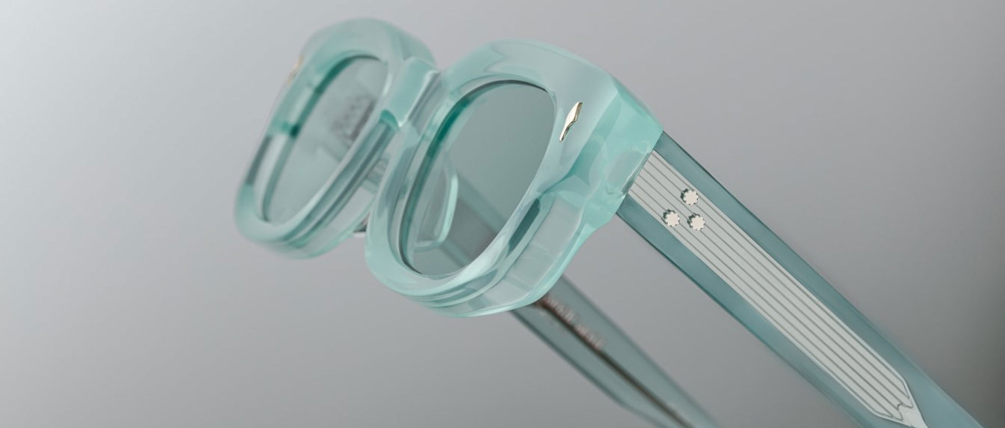 Close-up of Jacques Marie Mage Whiskeyclone eyeglass frames in translucent mint green with thick rims and two silver dots on the temples, set against a plain light grey background.