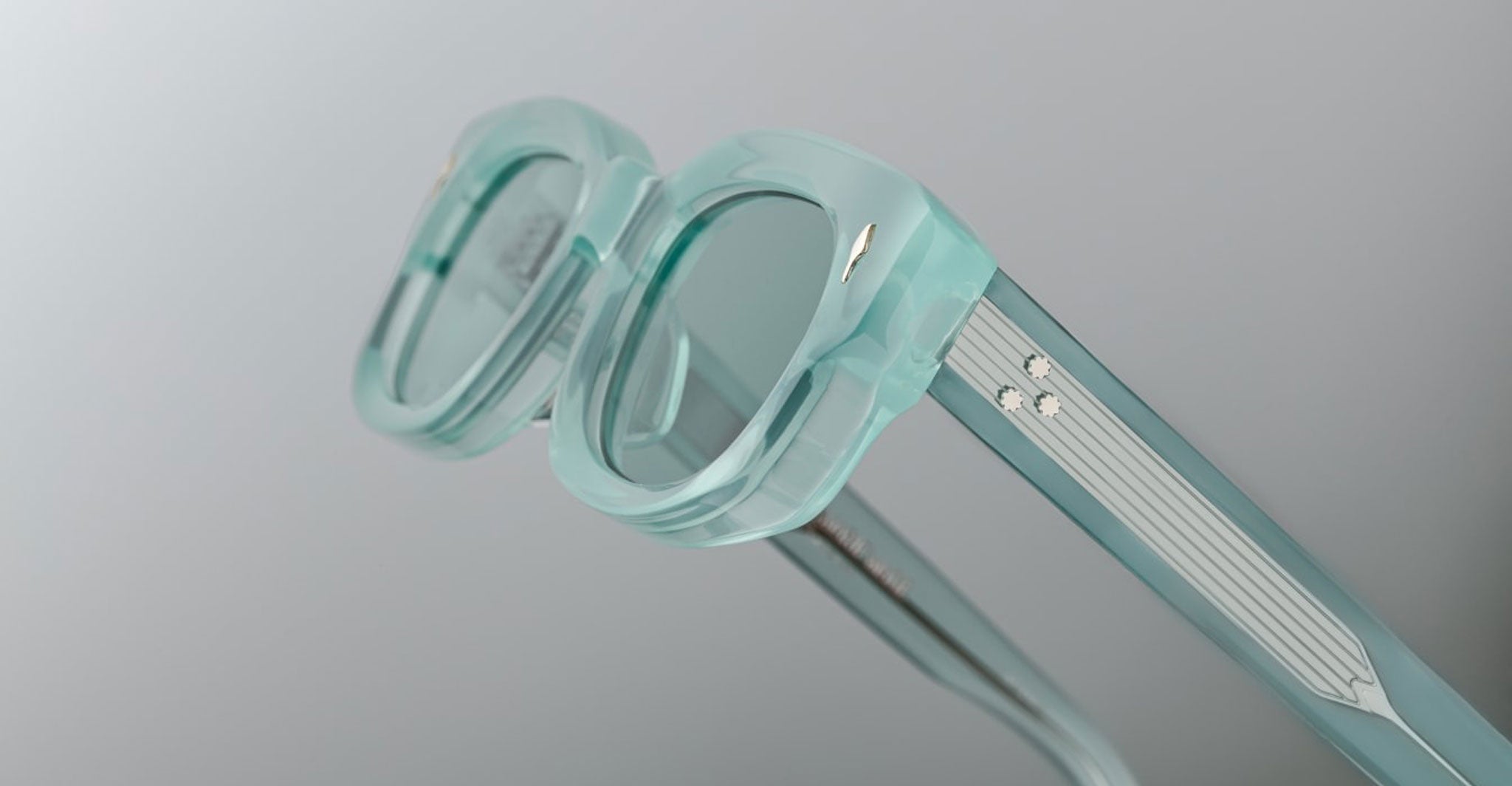 Close-up of Jacques Marie Mage Whiskeyclone eyeglass frames in translucent mint green with thick rims and two silver dots on the temples, set against a plain light grey background.
