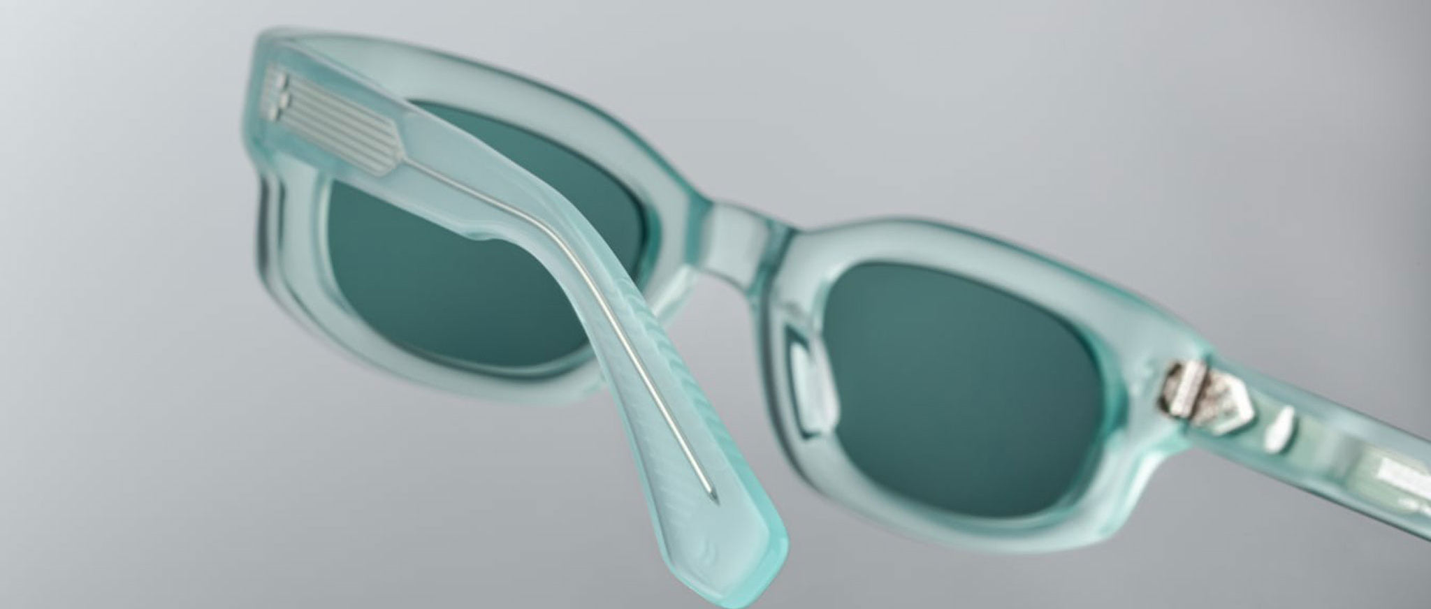 Close-up of Jacques Marie Mage Whiskeyclone sunglasses featuring translucent light blue frames and dark lenses, highlighting the frame and one arm against a plain grey background.