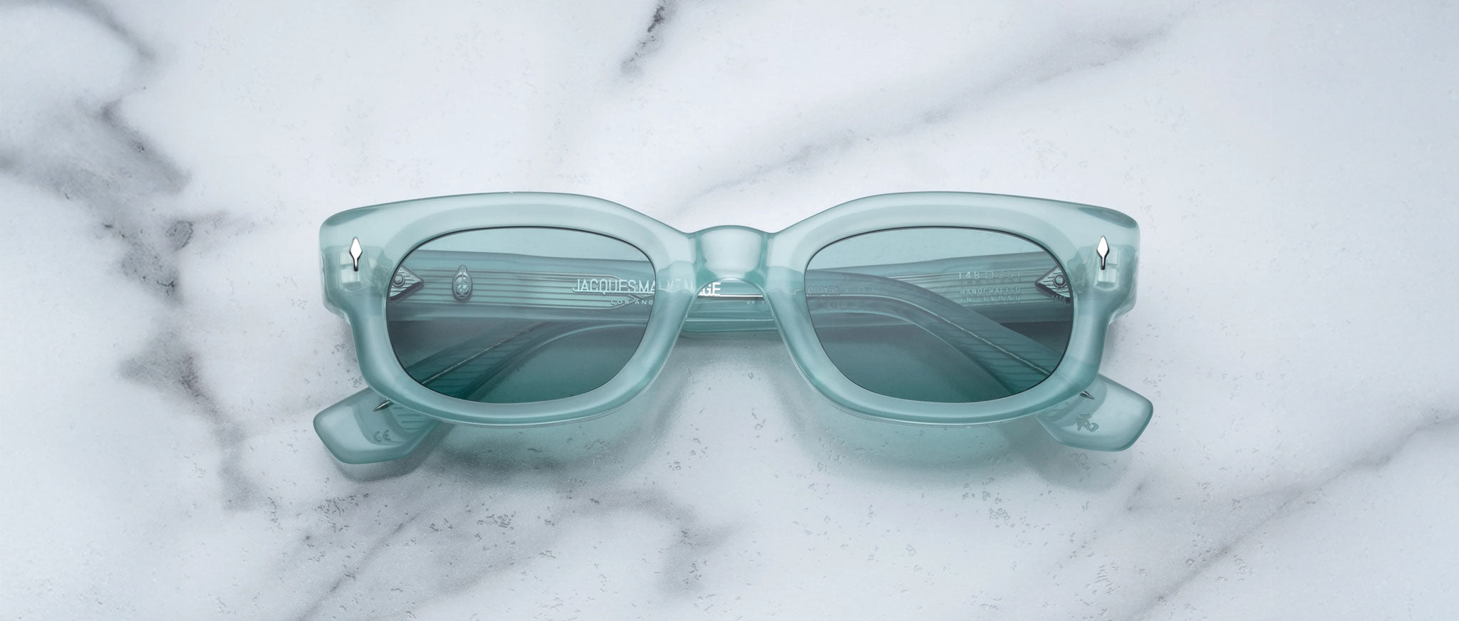 A pair of Jacques Marie Mage Whiskeyclone rectangular, light blue sunglasses with dark lenses rests on a white marble surface featuring grey veining.