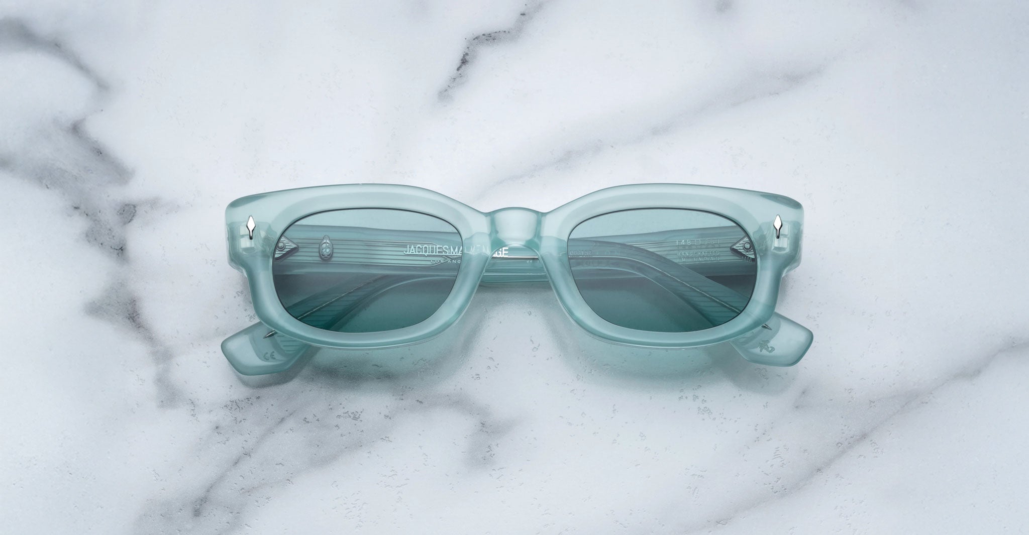 A pair of Jacques Marie Mage Whiskeyclone rectangular, light blue sunglasses with dark lenses rests on a white marble surface featuring grey veining.