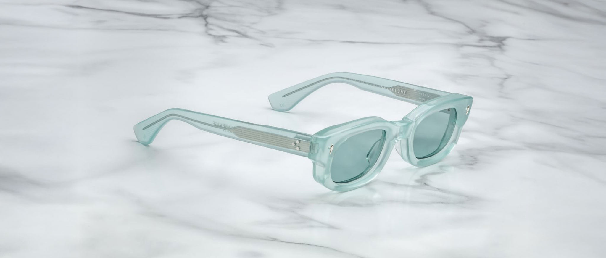 The Jacques Marie Mage Whiskeyclone sunglasses, in light blue translucent with thick frames and matching tinted lenses, are displayed on a white marble surface with gray veining.