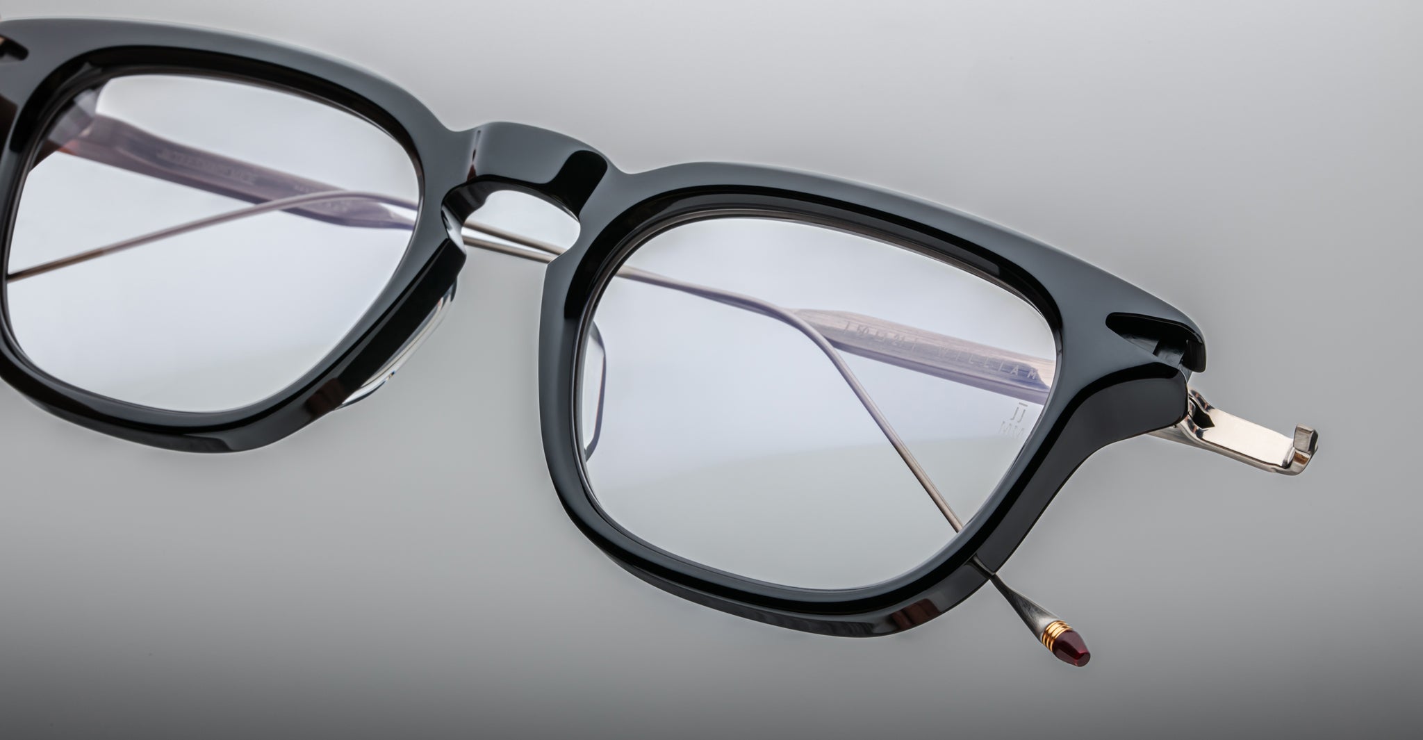 Close-up of Jacques Marie Mage William black rectangular eyeglasses with metal hinges and clear lenses, resting on a smooth light gray surface. The frame and temples are partially visible.
