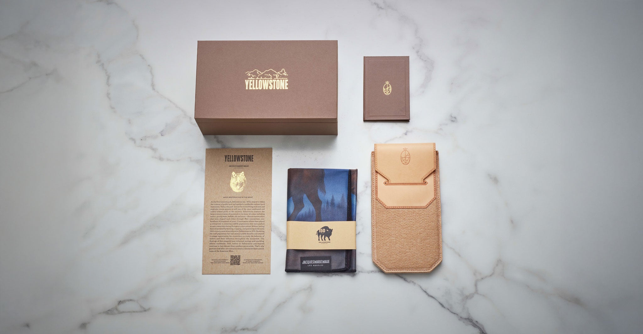 A Jacques Marie Mage Firehole River set—featuring a brown Yellowstone box, small brown booklet, tan leather pouch, card, and folded blue cloth with the Yellowstone bison logo—is displayed on white marble.
