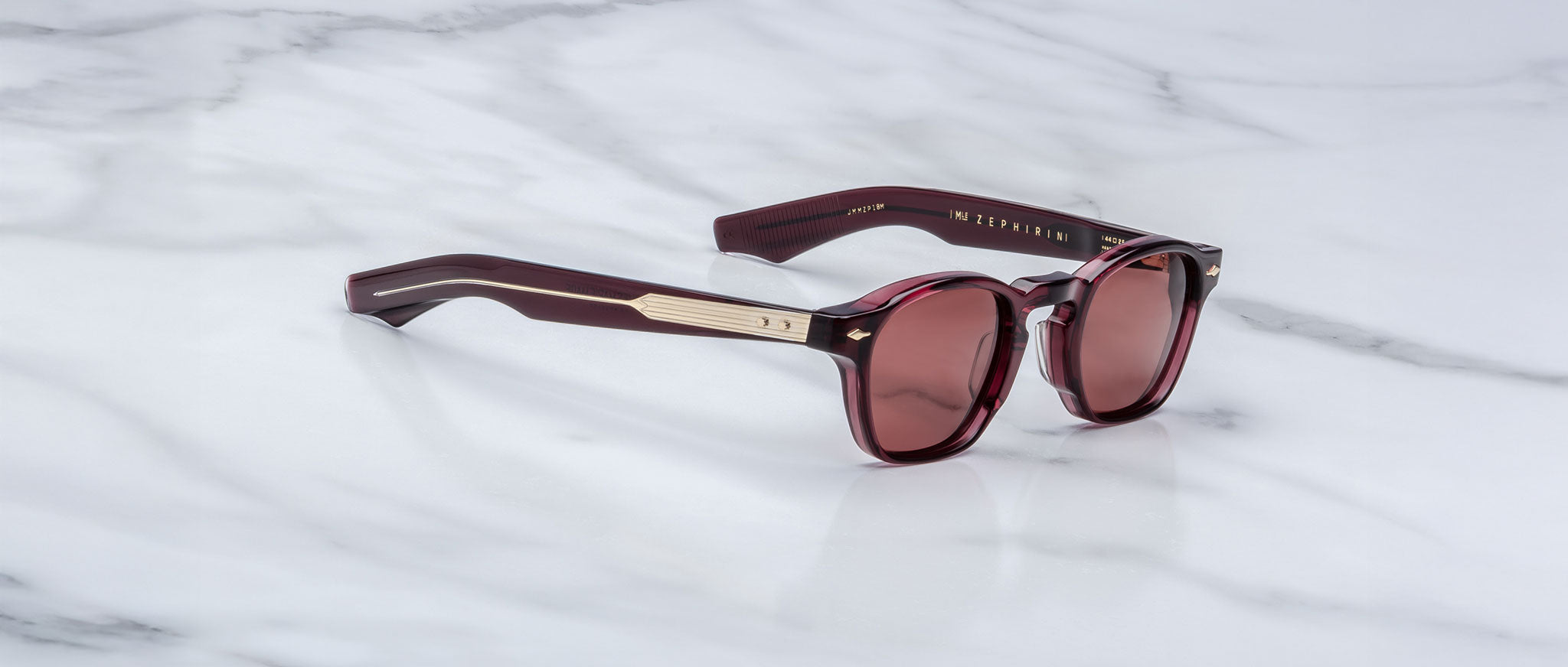 The Jacques Marie Mage Zephirin sunglasses feature maroon acetate frames, gold accents, and round tinted lenses. This handcrafted, limited-edition eyewear by Jacques Marie Mage rests elegantly on a white marble surface.