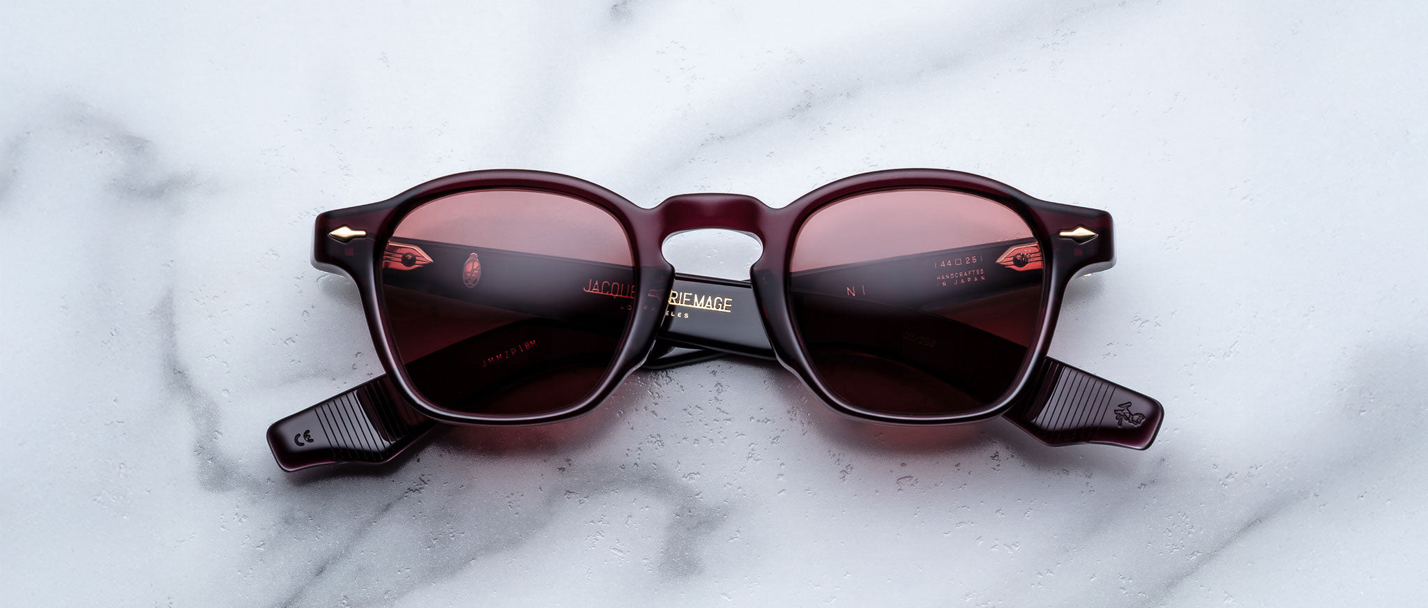 A pair of Jacques Marie Mage Zephirin sunglasses with round, burgundy frames and red-tinted lenses rests on a white marble surface—showcasing the brand’s exquisite limited edition eyewear.