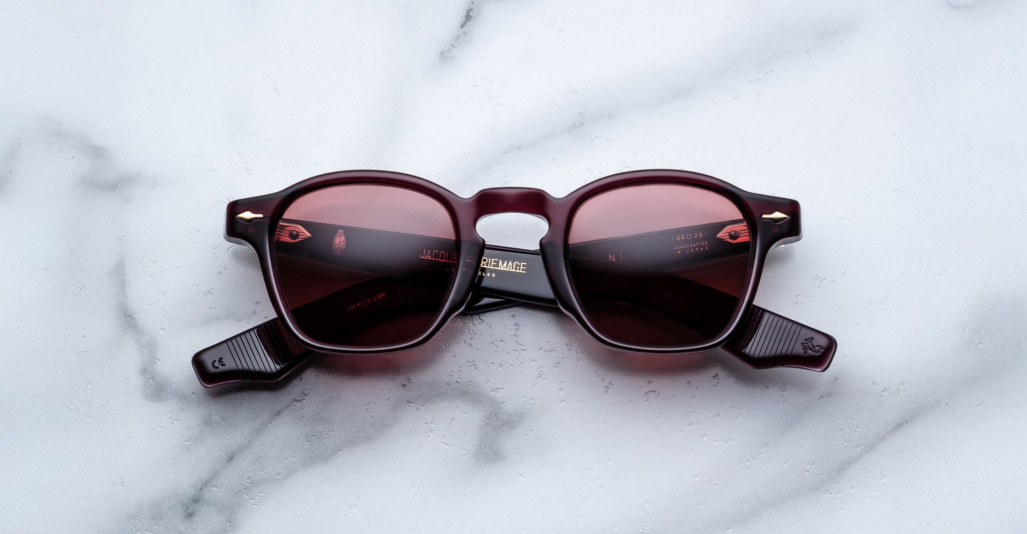 A pair of Jacques Marie Mage Zephirin sunglasses with round, burgundy frames and red-tinted lenses rests on a white marble surface—showcasing the brand’s exquisite limited edition eyewear.