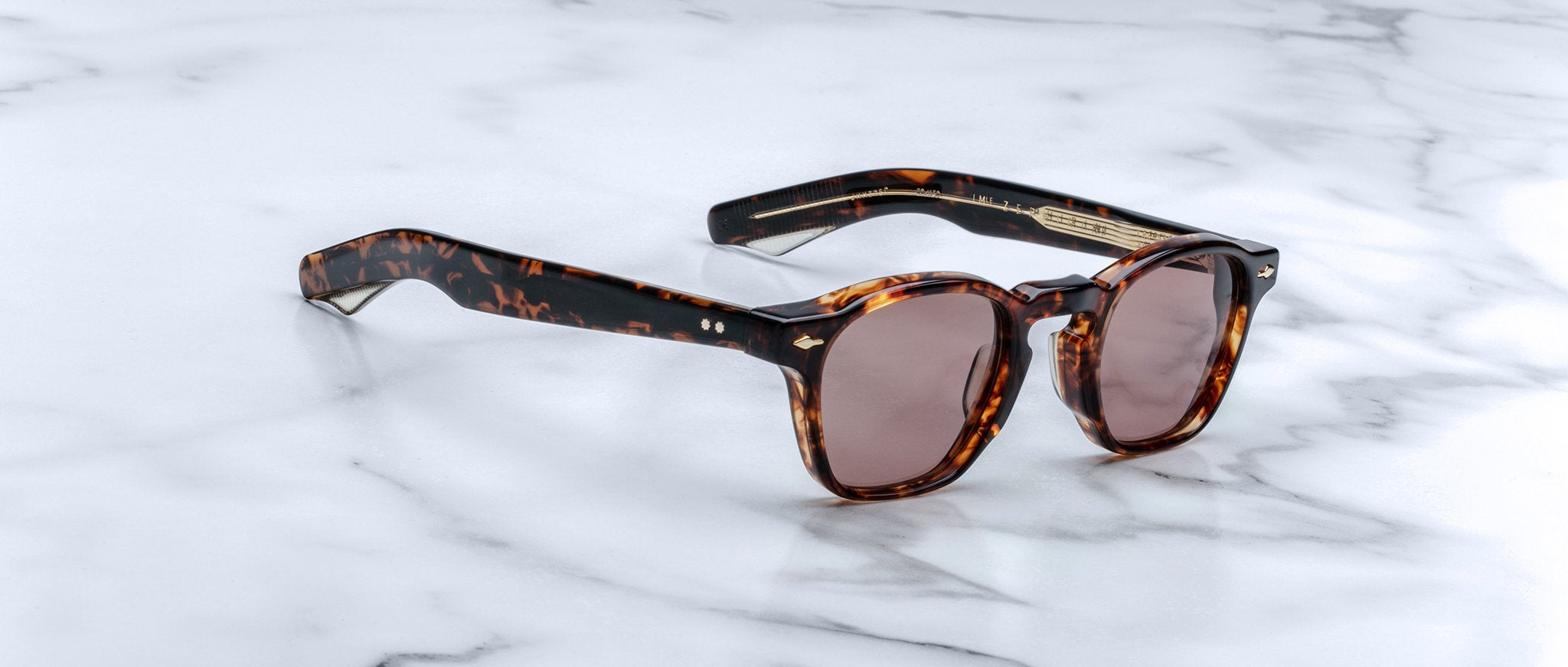 Limited edition Jacques Marie Mage Zephirin sunglasses, crafted from handcrafted acetate with a tortoiseshell pattern and round brown lenses, are displayed on a white marble surface.