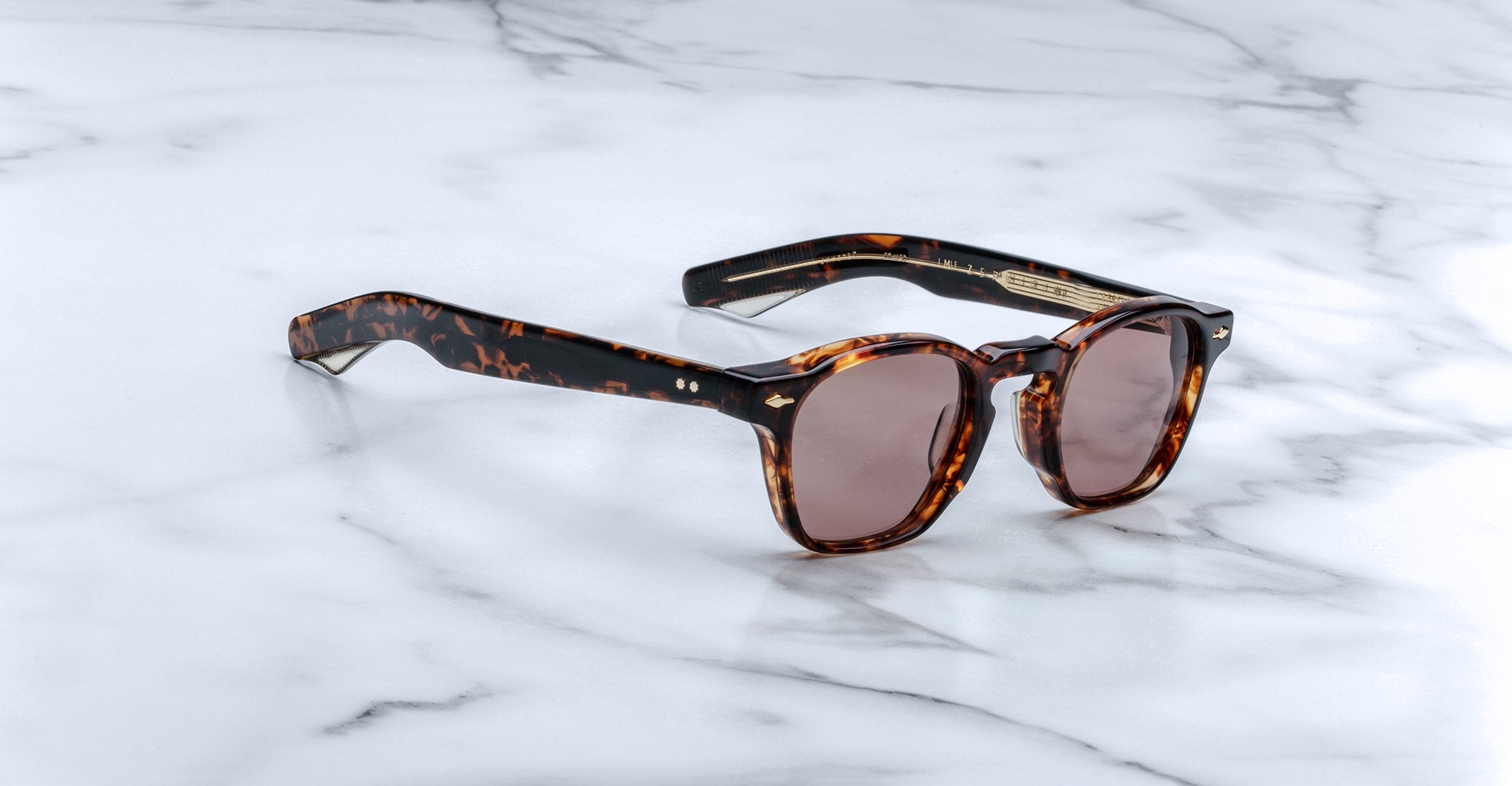 Limited edition Jacques Marie Mage Zephirin sunglasses, crafted from handcrafted acetate with a tortoiseshell pattern and round brown lenses, are displayed on a white marble surface.