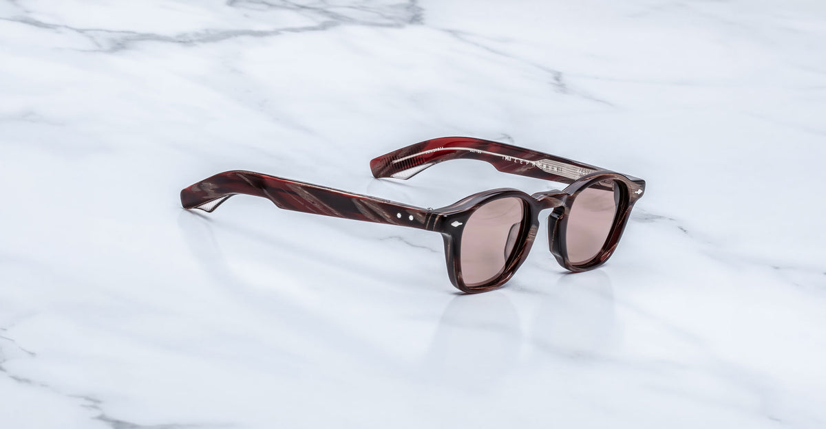 Jacques Marie Mage Zephirin sunglasses in Auburn, three-quarter view with reddish-brown marbled acetate, rose-tinted lenses, and silver-tone accents on a white marble surface.