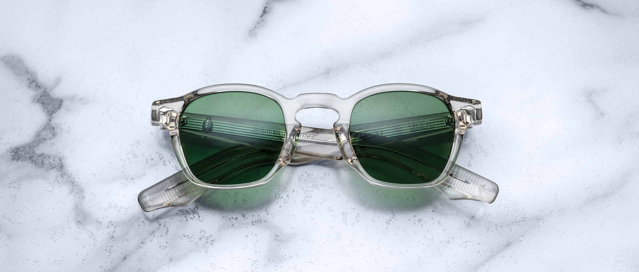 Jacques Marie Mage Zephirin sunglasses by Jacques Marie Mage, featuring clear frames and green lenses inspired by handcrafted acetate spectacles, are displayed folded on a white marble surface.