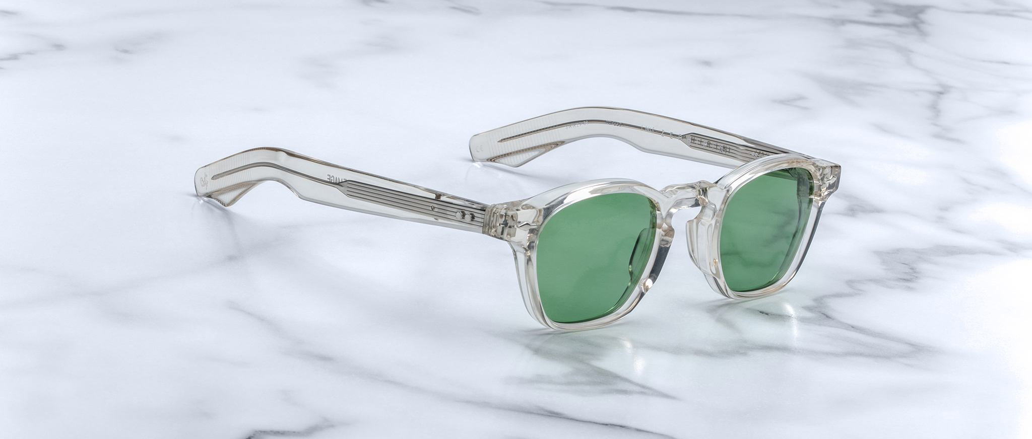 Jacques Marie Mage Zephirin sunglasses with clear acetate frames and green lenses by Jacques Marie Mage are displayed on a white marble surface.