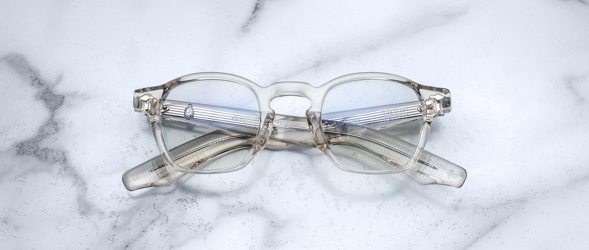 The Jacques Marie Mage Zephirin eyeglasses by Jacques Marie Mage, featuring round lenses and translucent frames, are folded and set on a white marble surface.