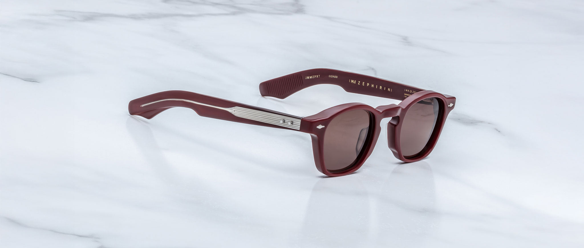 The Jacques Marie Mage Zephirin sunglasses by Jacques Marie Mage feature round maroon lenses, metallic arm accents, and a limited edition-inspired design, elegantly displayed on a white marble surface.