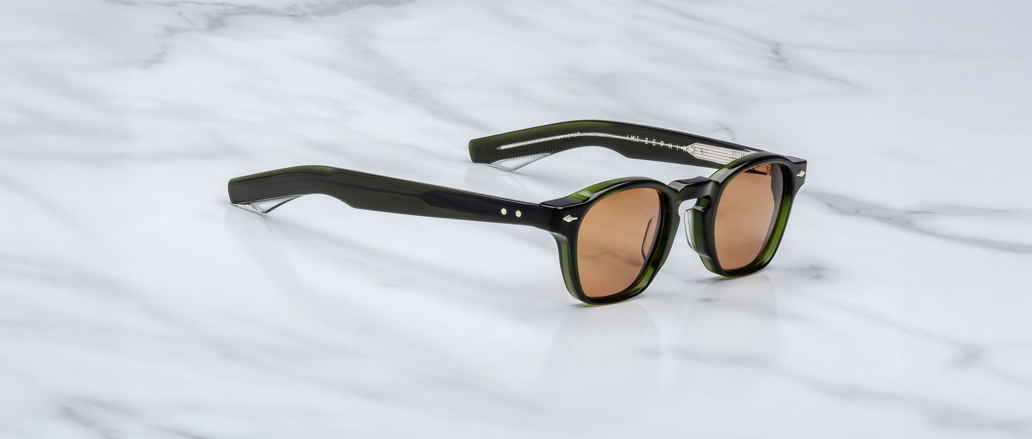 A pair of limited edition Jacques Marie Mage Zephirin sunglasses in dark green with brown lenses, crafted from premium acetate, rests on a white marble surface.