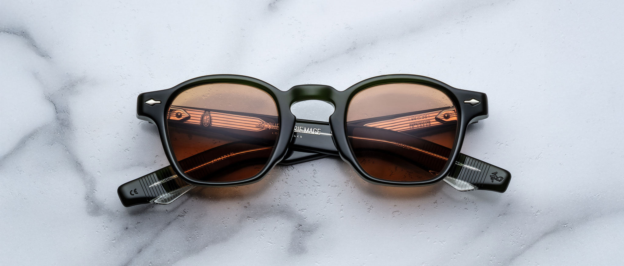 A pair of Jacques Marie Mage Zephirin sunglasses with round dark frames and brown-tinted lenses rests on white marble, highlighting the charm of this limited edition Jacques Marie Mage eyewear.