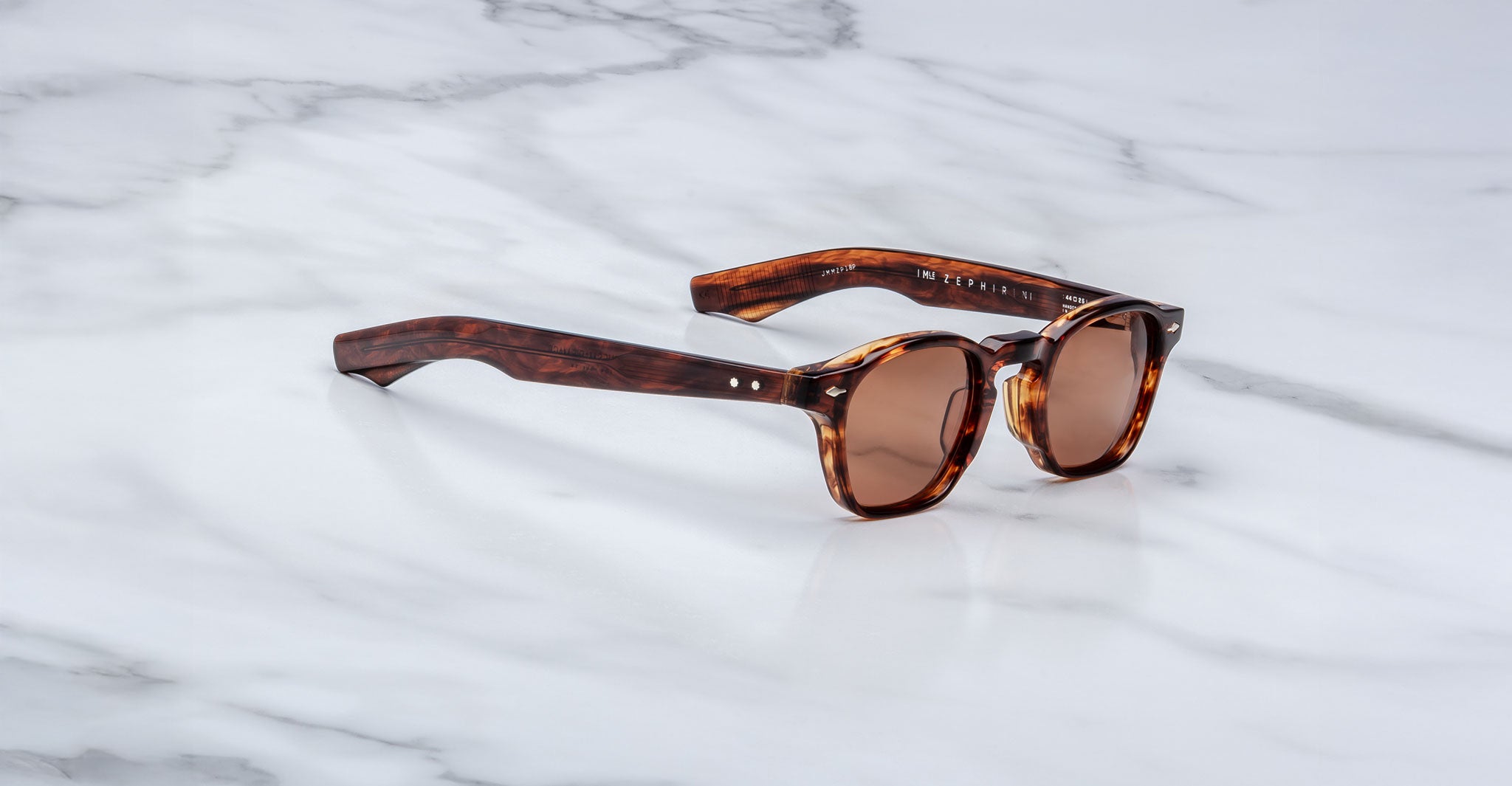 A handcrafted pair of Jacques Marie Mage Zephirin sunglasses featuring brown tortoiseshell acetate, round lenses, and thick frames, elegantly displayed on a white marble surface.