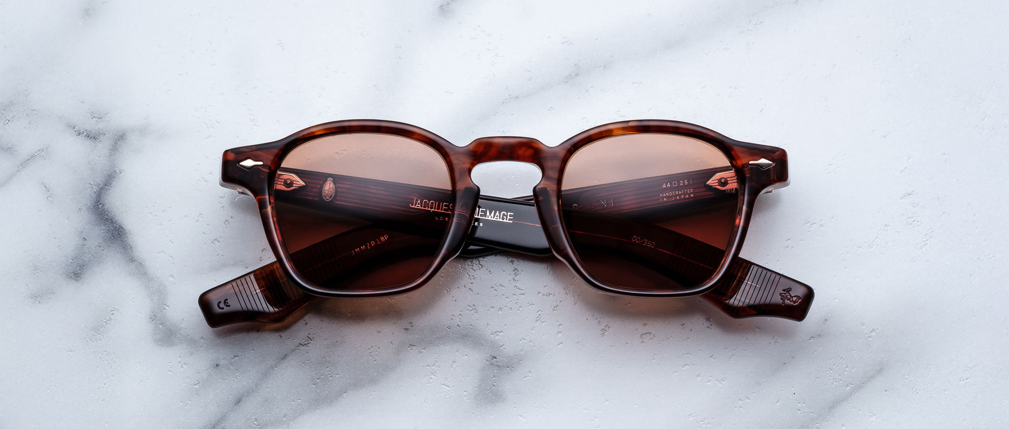 The Jacques Marie Mage Zephirin sunglasses by Jacques Marie Mage feature a brown tortoiseshell frame with red-tinted lenses, inspired by handcrafted acetate designs, displayed on a white marble surface.