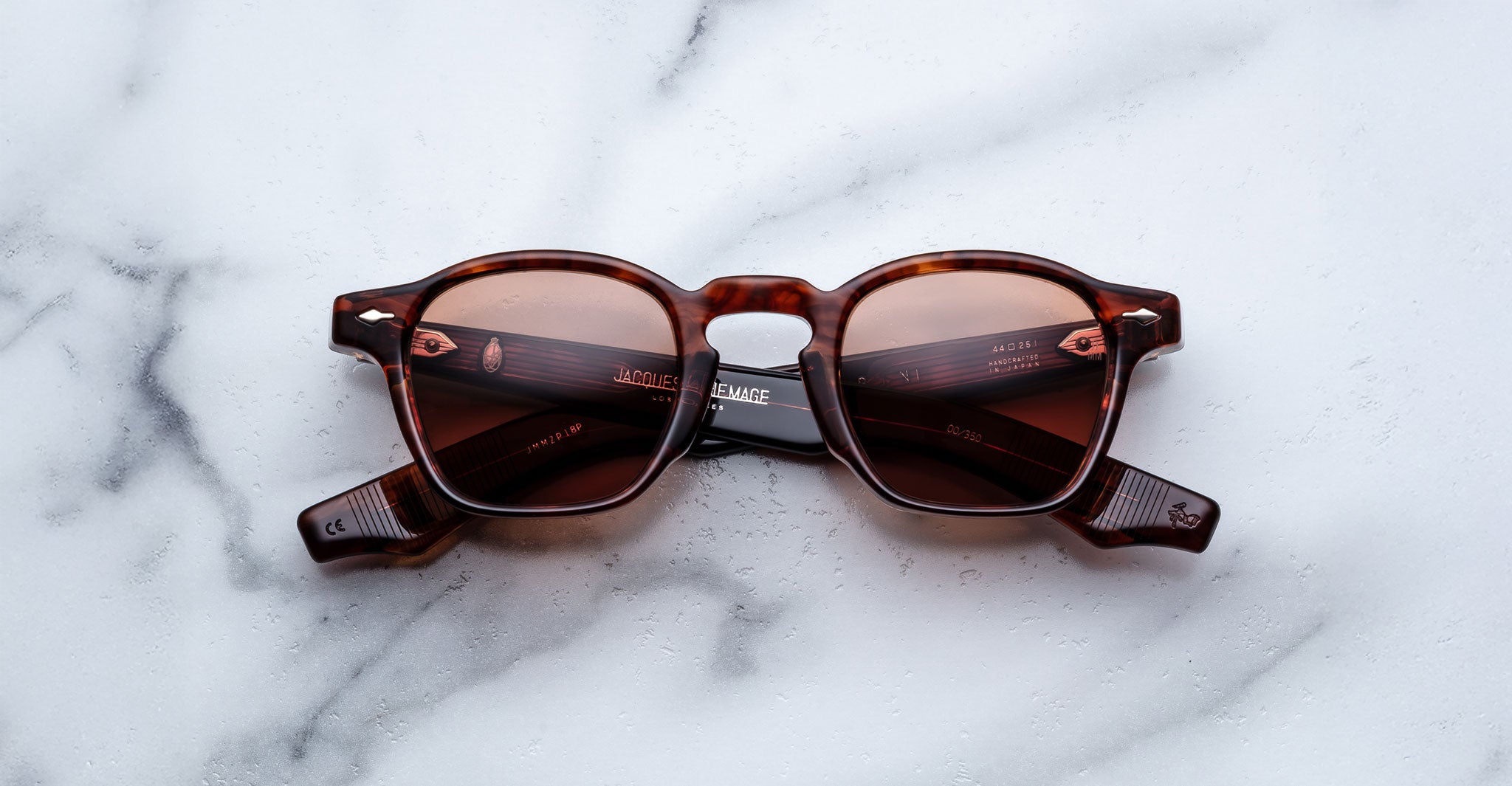 The Jacques Marie Mage Zephirin sunglasses by Jacques Marie Mage feature a brown tortoiseshell frame with red-tinted lenses, inspired by handcrafted acetate designs, displayed on a white marble surface.