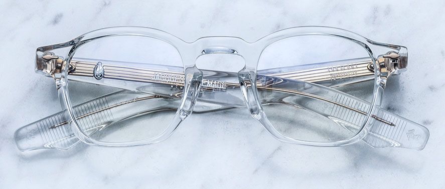 Jacques Marie Mage Zephirin spectacles by Jacques Marie Mage feature clear handcrafted acetate frames, rectangular lenses, and metal hinges. Temples crossed on a white marble surface—a subtle nod to limited edition eyewear.