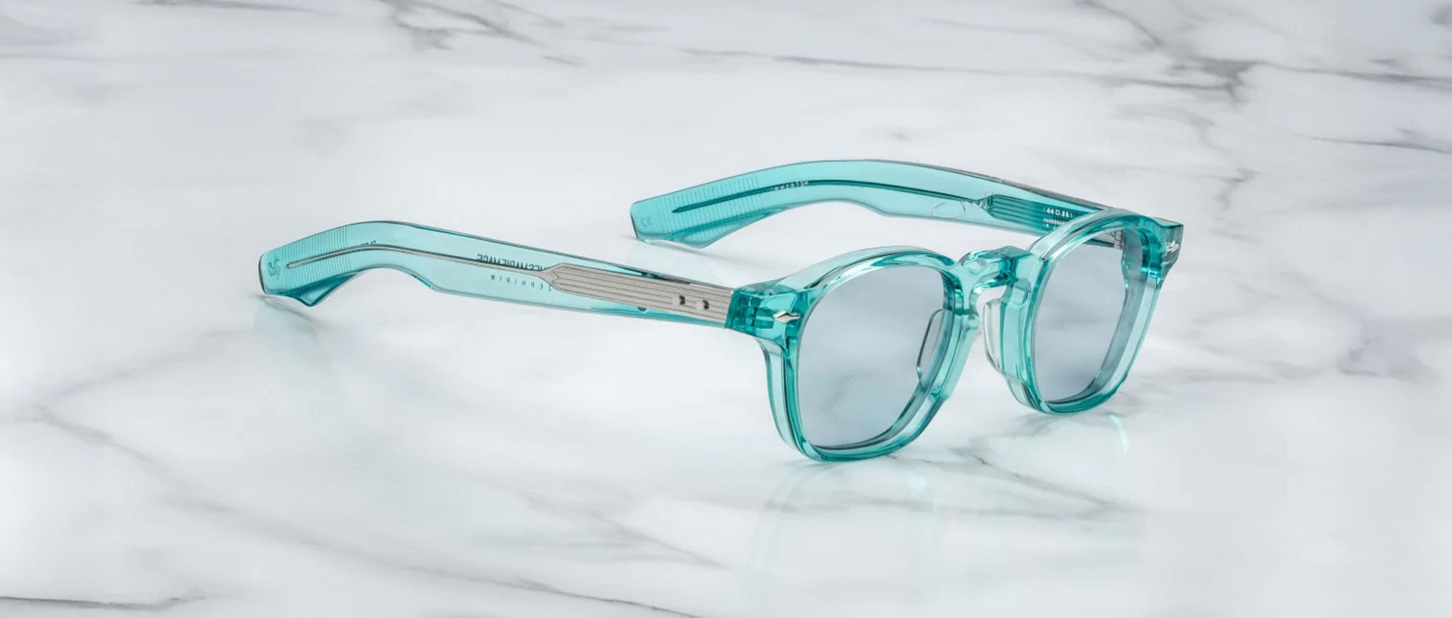 The Jacques Marie Mage Zephirin by Jacques Marie Mage features handcrafted acetate frames in transparent teal with round, thick lenses, elegantly displayed on white marble—a sophisticated limited edition eyewear piece.