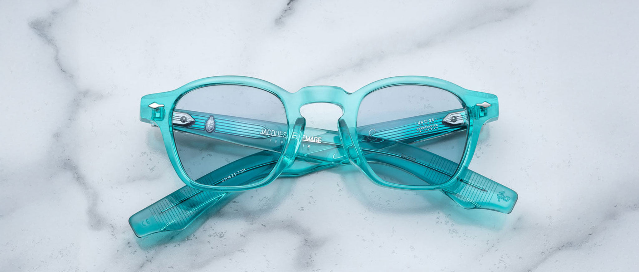 A pair of Jacques Marie Mage Zephirin turquoise translucent handcrafted acetate spectacles with clear lenses rests closed on a white marble surface.