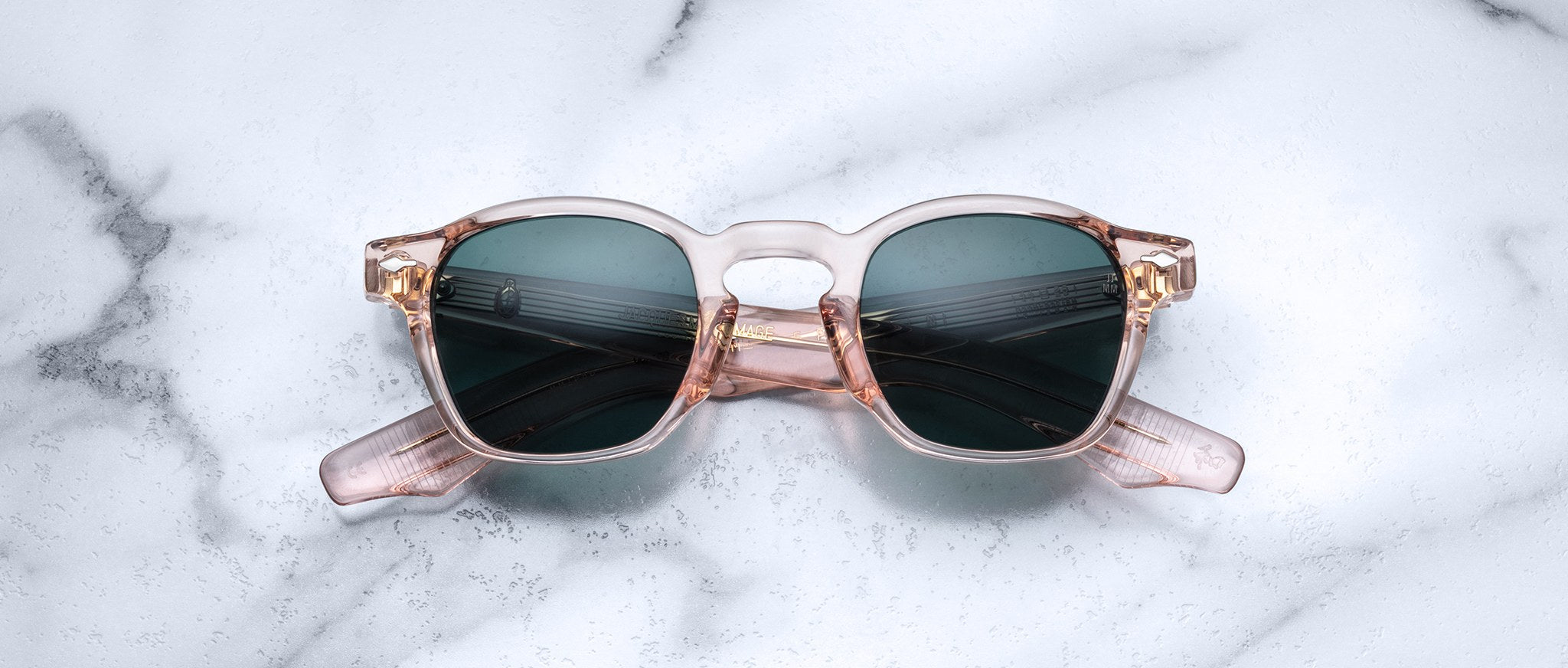 The Jacques Marie Mage Zephirin by Jacques Marie Mage features transparent pink handcrafted acetate frames with dark lenses, displayed on a white marble surface.