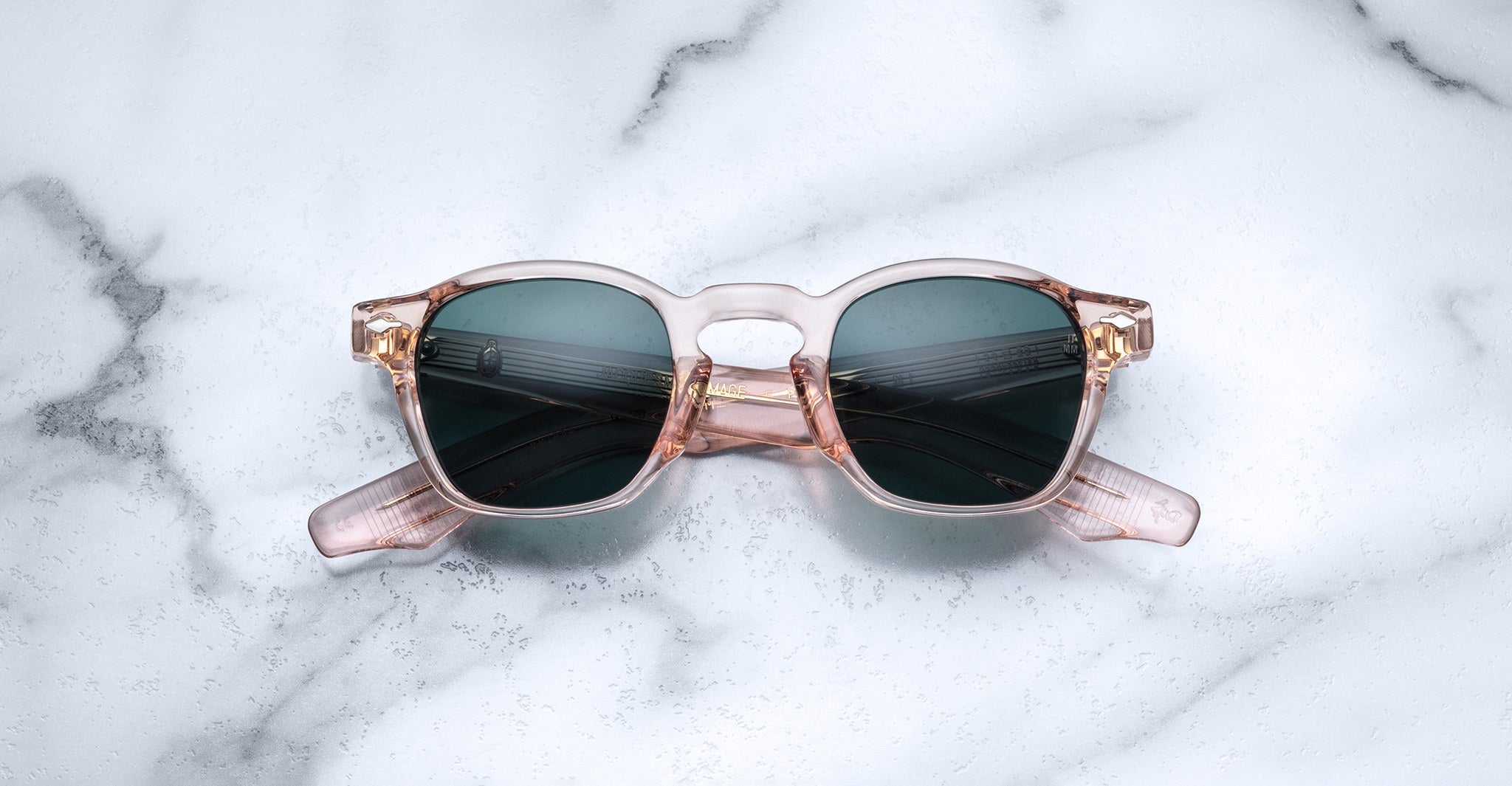 The Jacques Marie Mage Zephirin by Jacques Marie Mage features transparent pink handcrafted acetate frames with dark lenses, displayed on a white marble surface.
