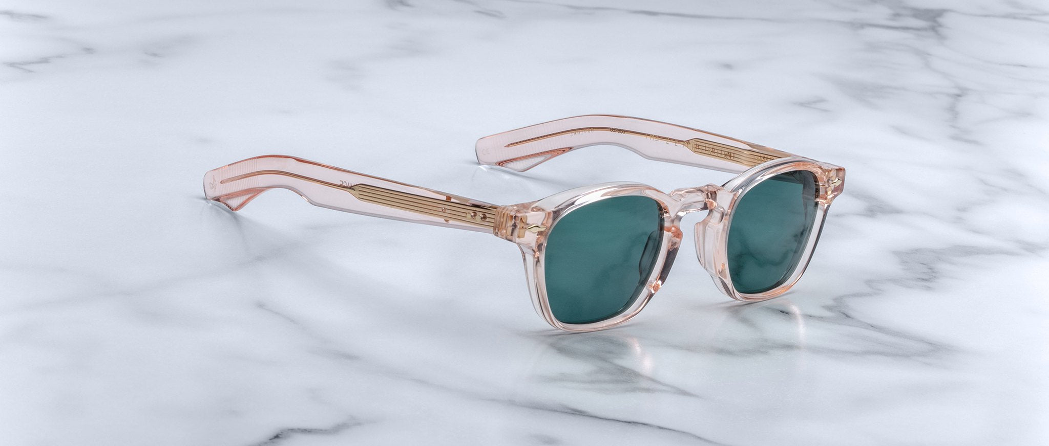 A pair of handcrafted Jacques Marie Mage Zephirin sunglasses in translucent pink with green lenses rest on a white marble surface, showcasing their limited edition appeal.