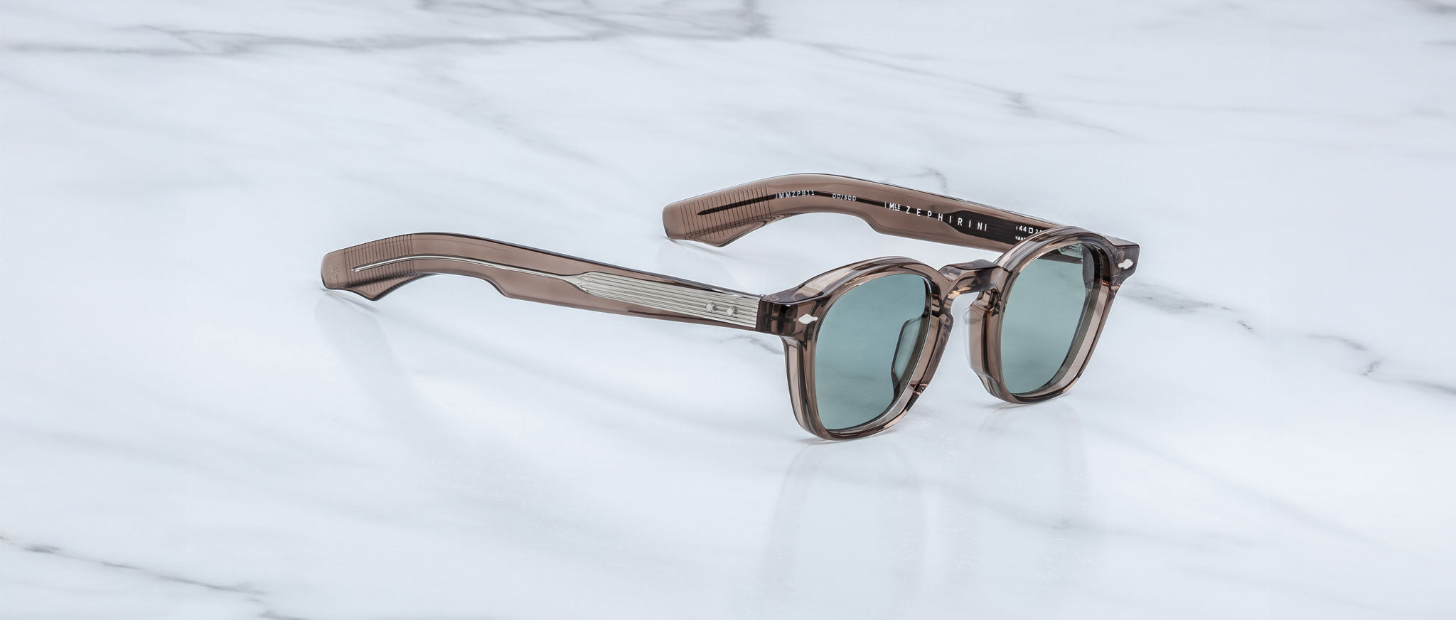 A pair of Jacques Marie Mage Zephirin sunglasses with brown translucent handcrafted acetate frames and round green lenses rests on a white marble surface.